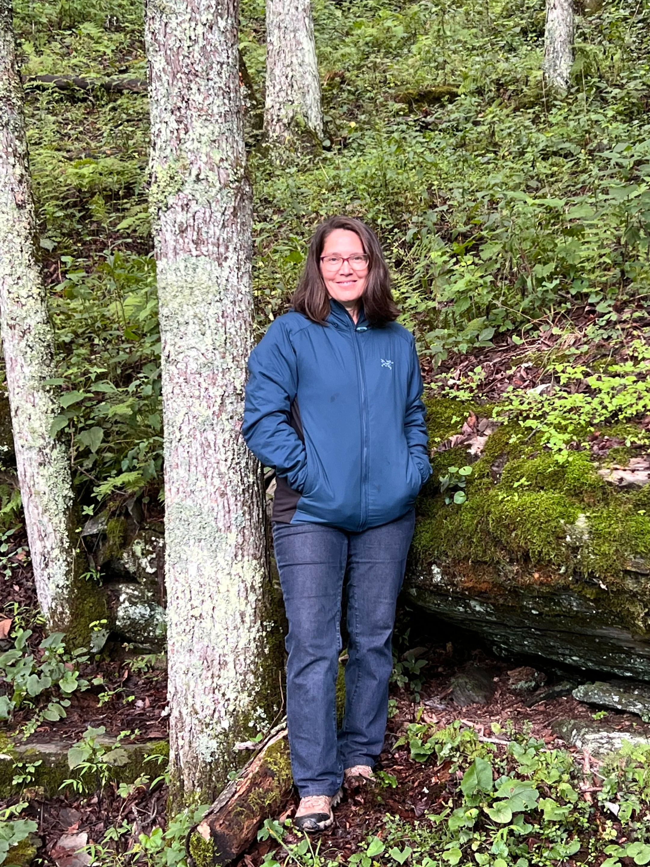 A woman with glasses and long dark hair smiling while standing in a lush green forest, wearing a blue jacket and dark pants.