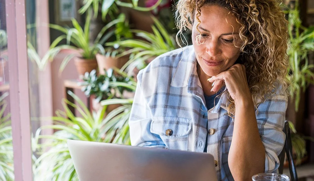 Woman wearing light blue shirt with curly hair sitting outside looking at laptop