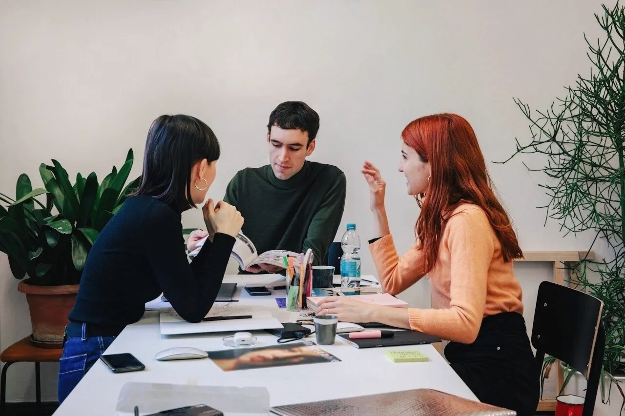Professional man in green jumper sitting in meeting with two women, one with a black polo neck and one with an orange blouse, collaborating in modern white office, with pens and cups on the white table and green plant in background