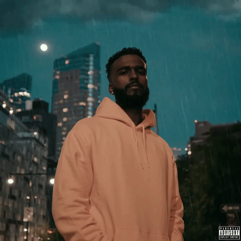 A man with a beard and short curly hair wearing a peach-colored hoodie stands outdoors in the rain at night, with tall buildings and the moon visible in the cloudy sky in the background.