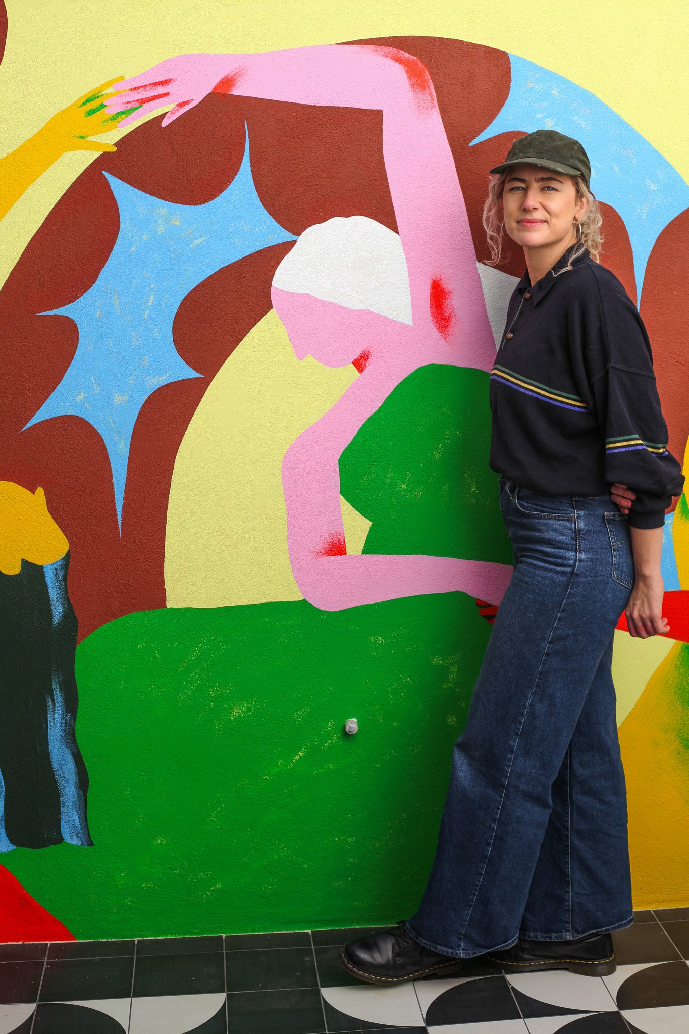 A woman with blonde standing beside a colorful abstract mural depicting a stylized female figure in pink, blue, green, and brown. Manonina is the artist, her name is Manon Nina. The mural is located in Sicily for a female co-living space.