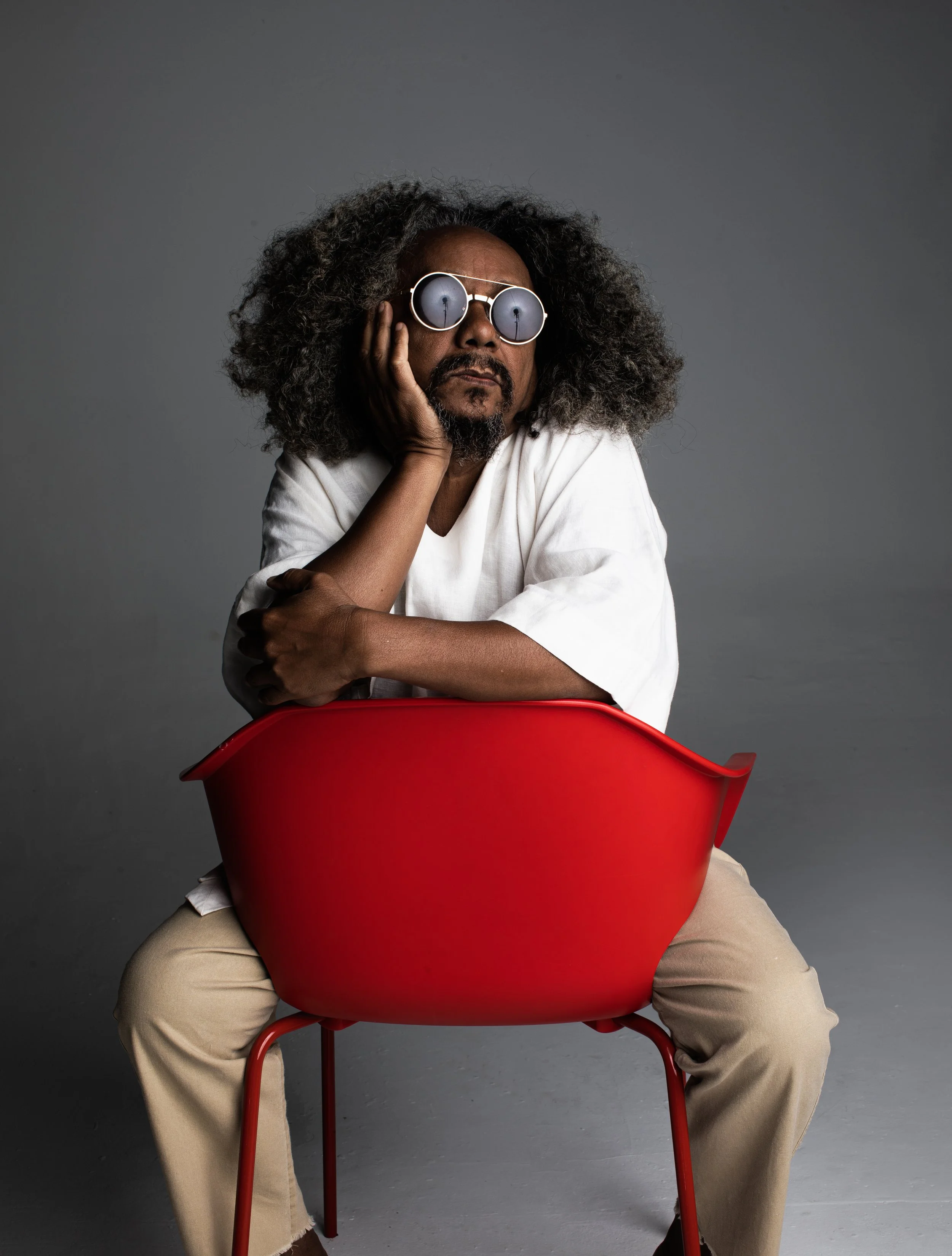 Homem com cabelo cacheado e óculos espelhados, sentado em uma cadeira vermelha, apoiando a cabeça na mão, vestindo camisa branca e calça bege.