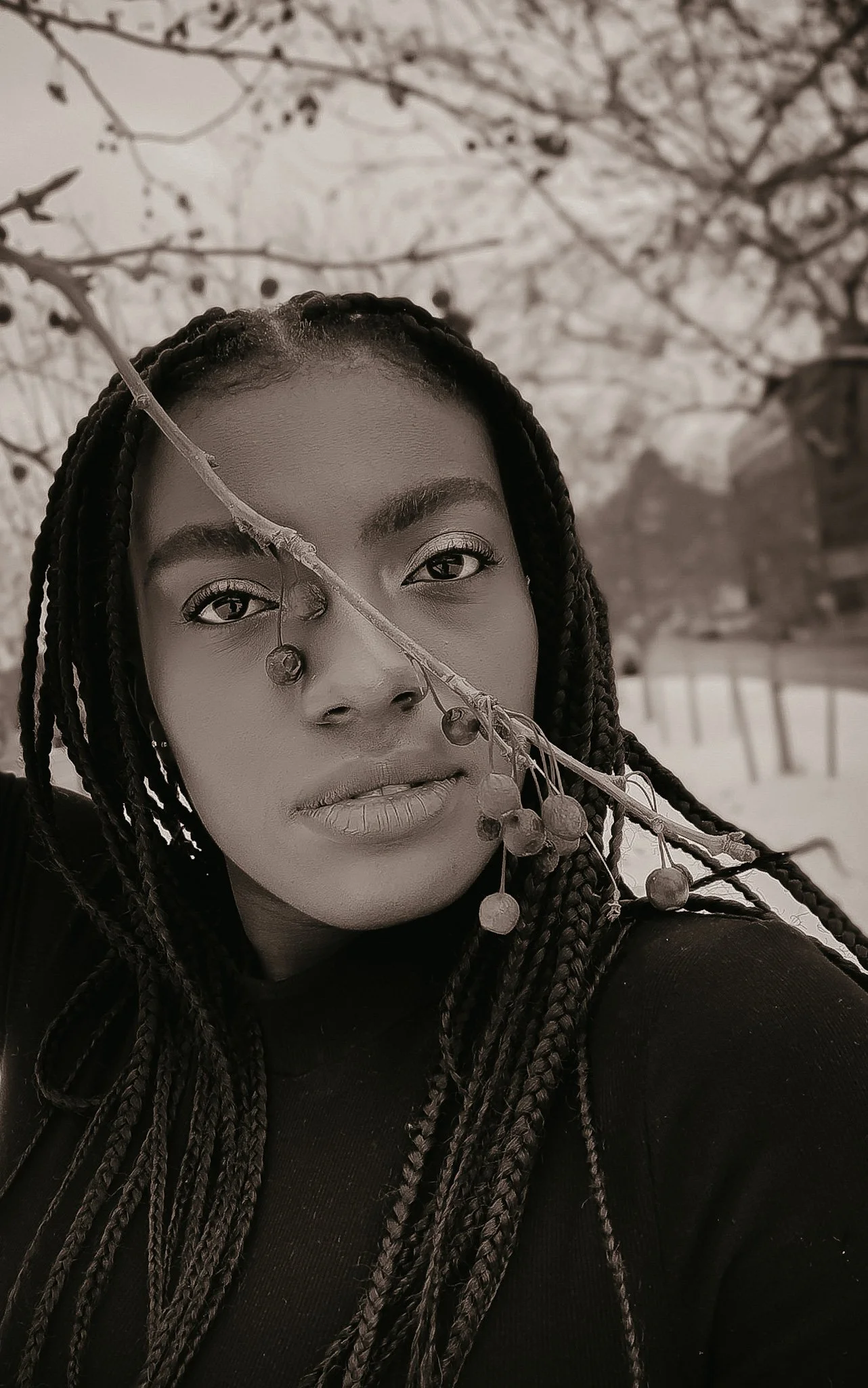 A black-and-white close-up portrait of a young woman with braided hair, standing outdoors near a tree with bare branches.