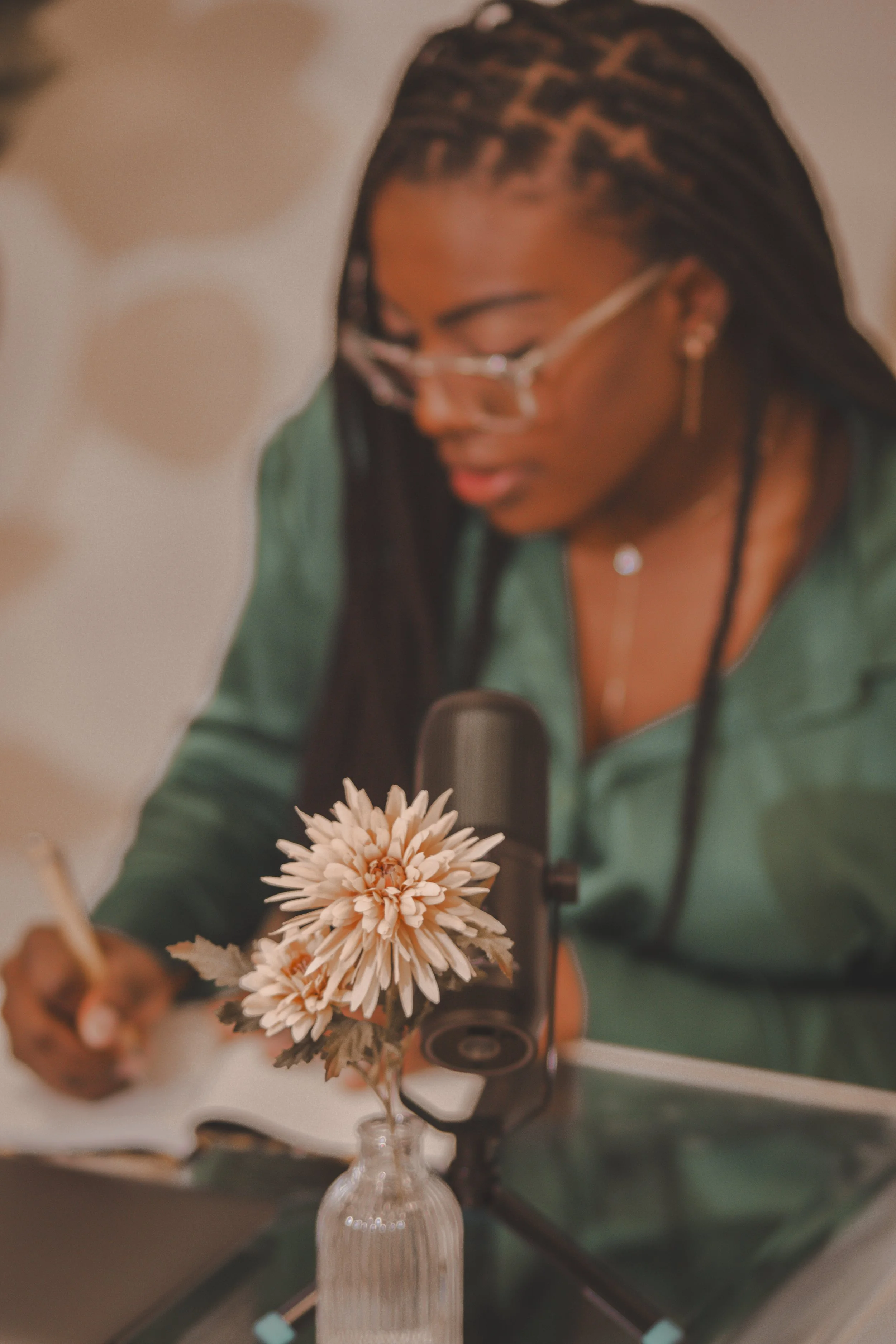 A woman with long braided hair and glasses is looking down, with a flower in a vase in the foreground. The background is blurred.