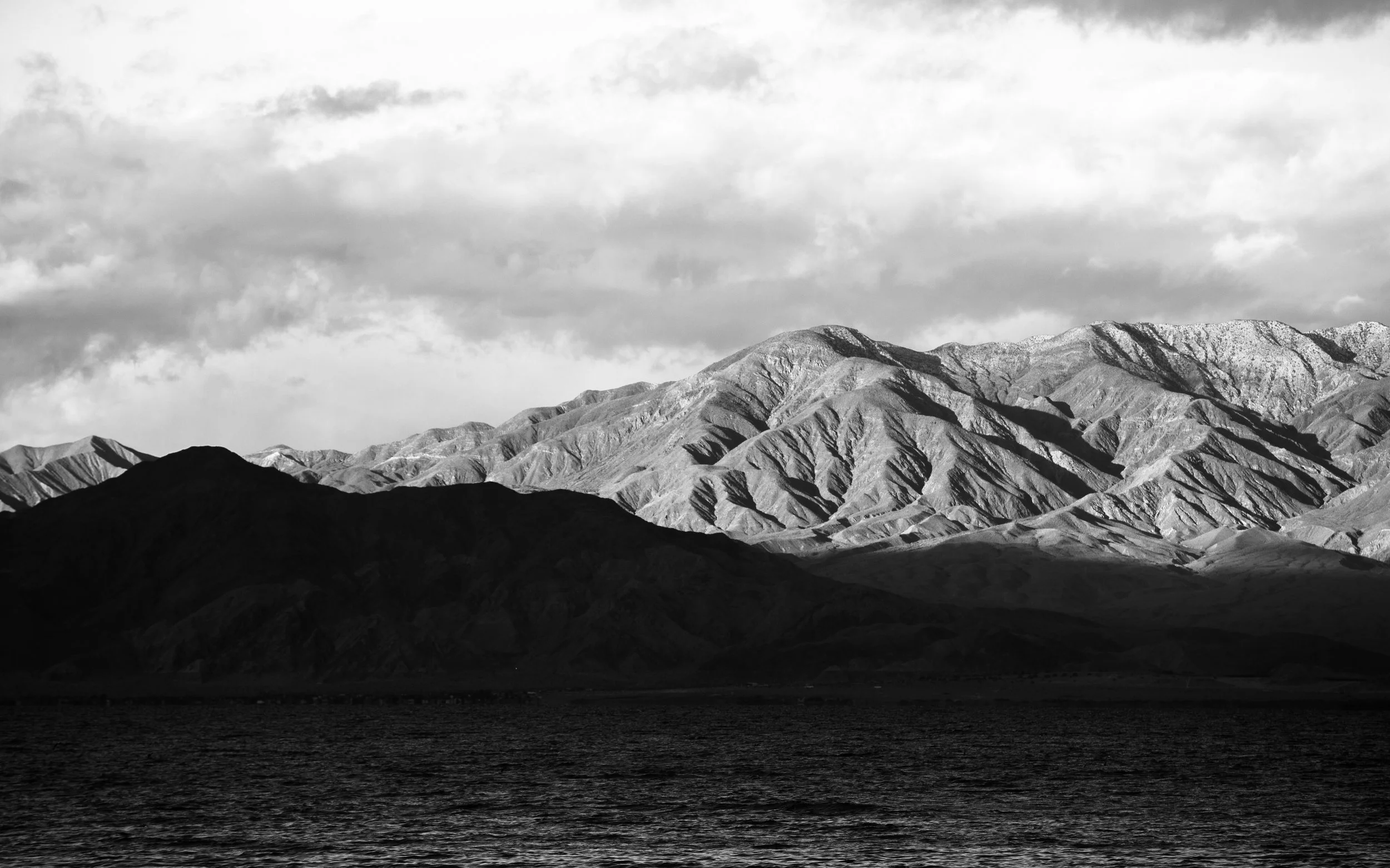 Black and white photograph of a mountain range with a body of water in the foreground. The mountains have varying shades of gray, and the sky above is partly cloudy.