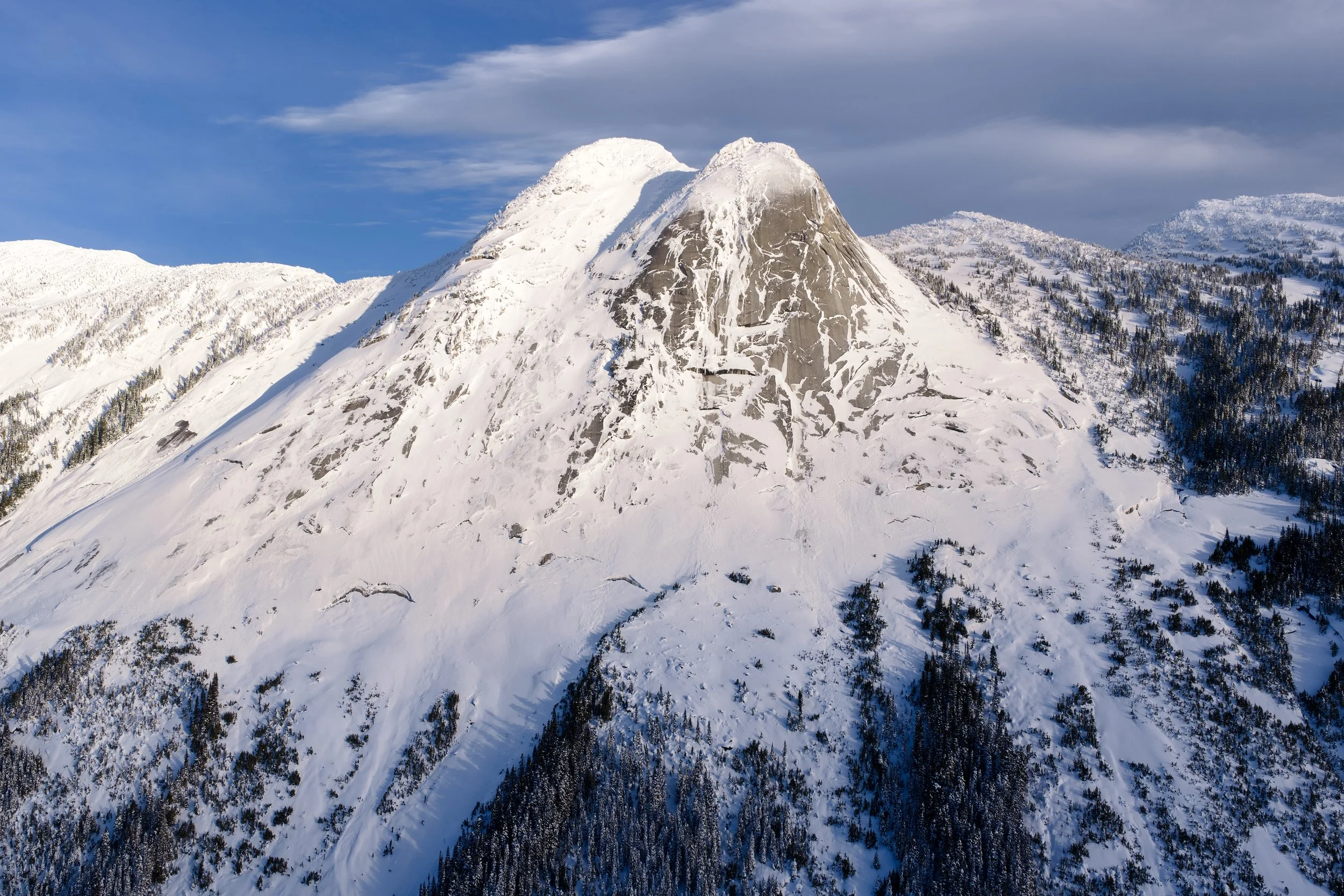 Snow-covered mountain with rocky face, surrounded by snowy slopes and pine trees, under a blue sky with clouds.