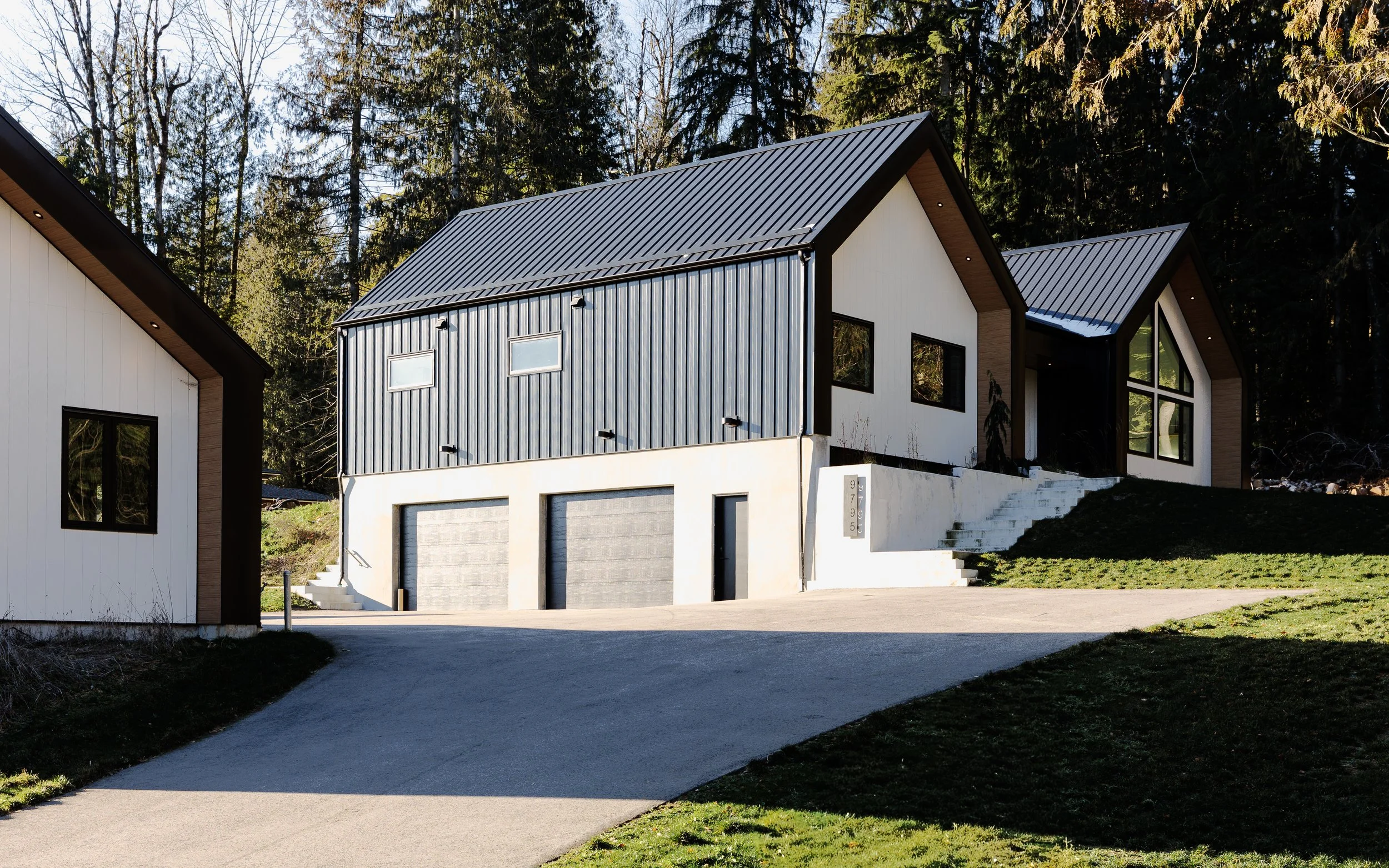 Modern house with black and white exterior, two-car garage, stairs leading up to the entrance, surrounded by trees and greenery.