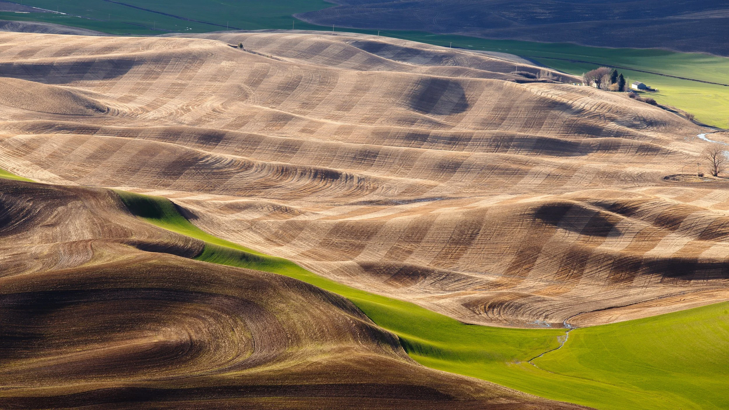 Aerial view of rolling hills with a mix of brown, green, and beige farmland, some with visible plowing patterns and a few small buildings in the distance.