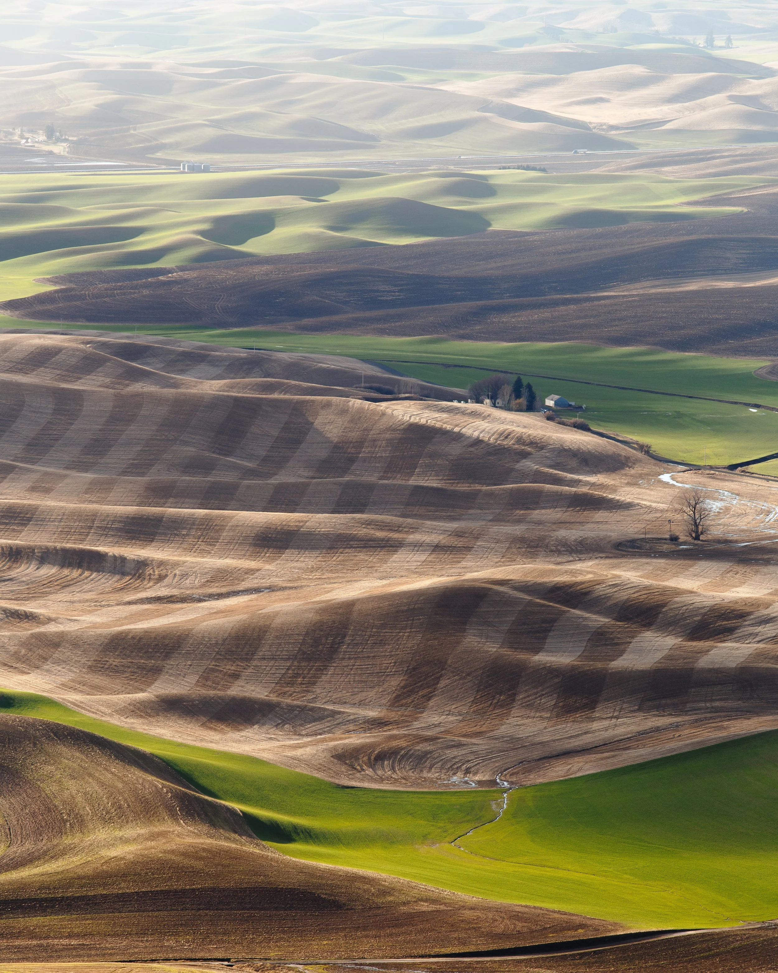Aerial view of rolling hills with patches of green grass and tilled farmland, some with trees and a few small buildings.