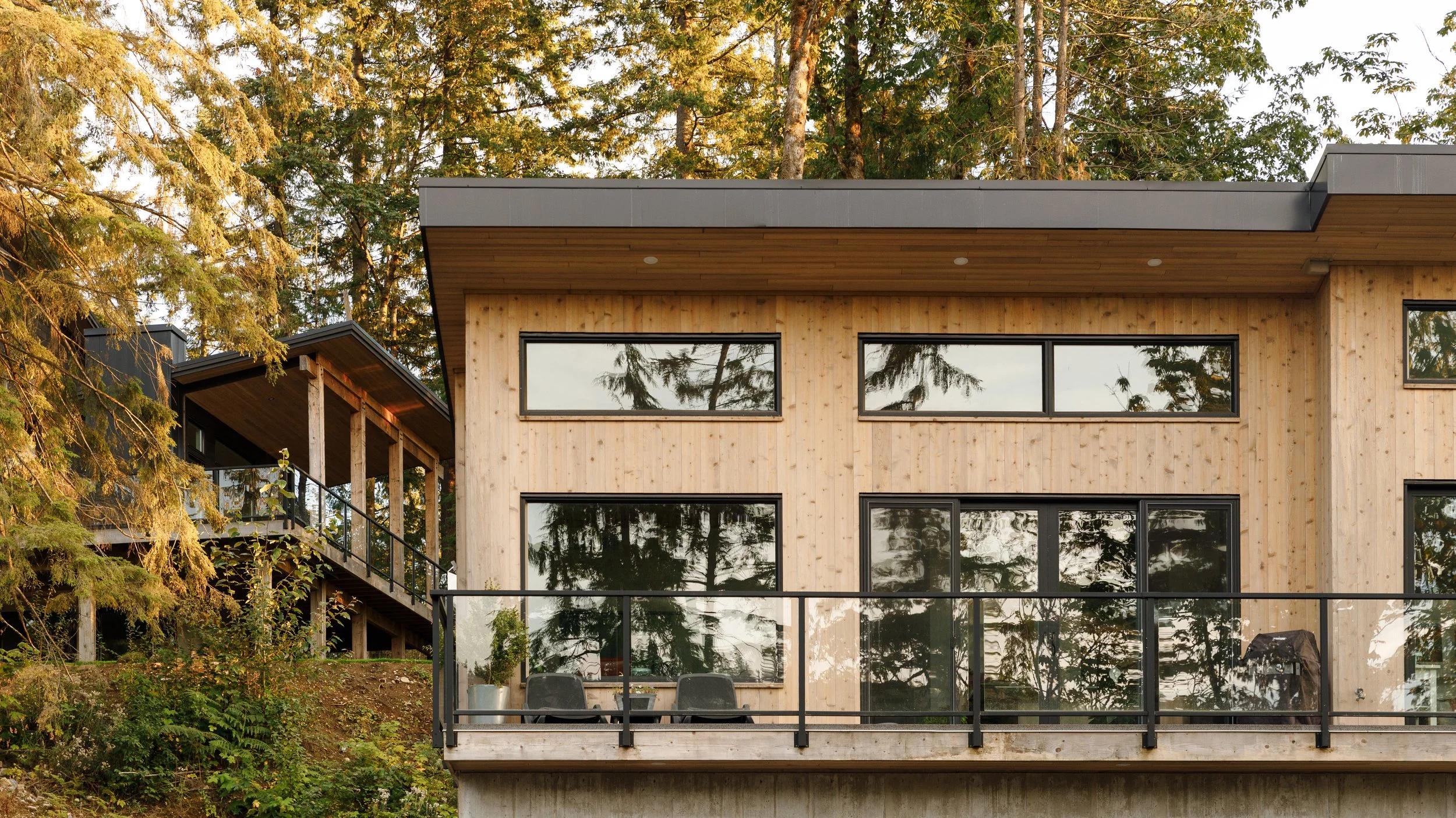 Modern two-story house with large windows, wooden exterior, and balcony surrounded by trees.
