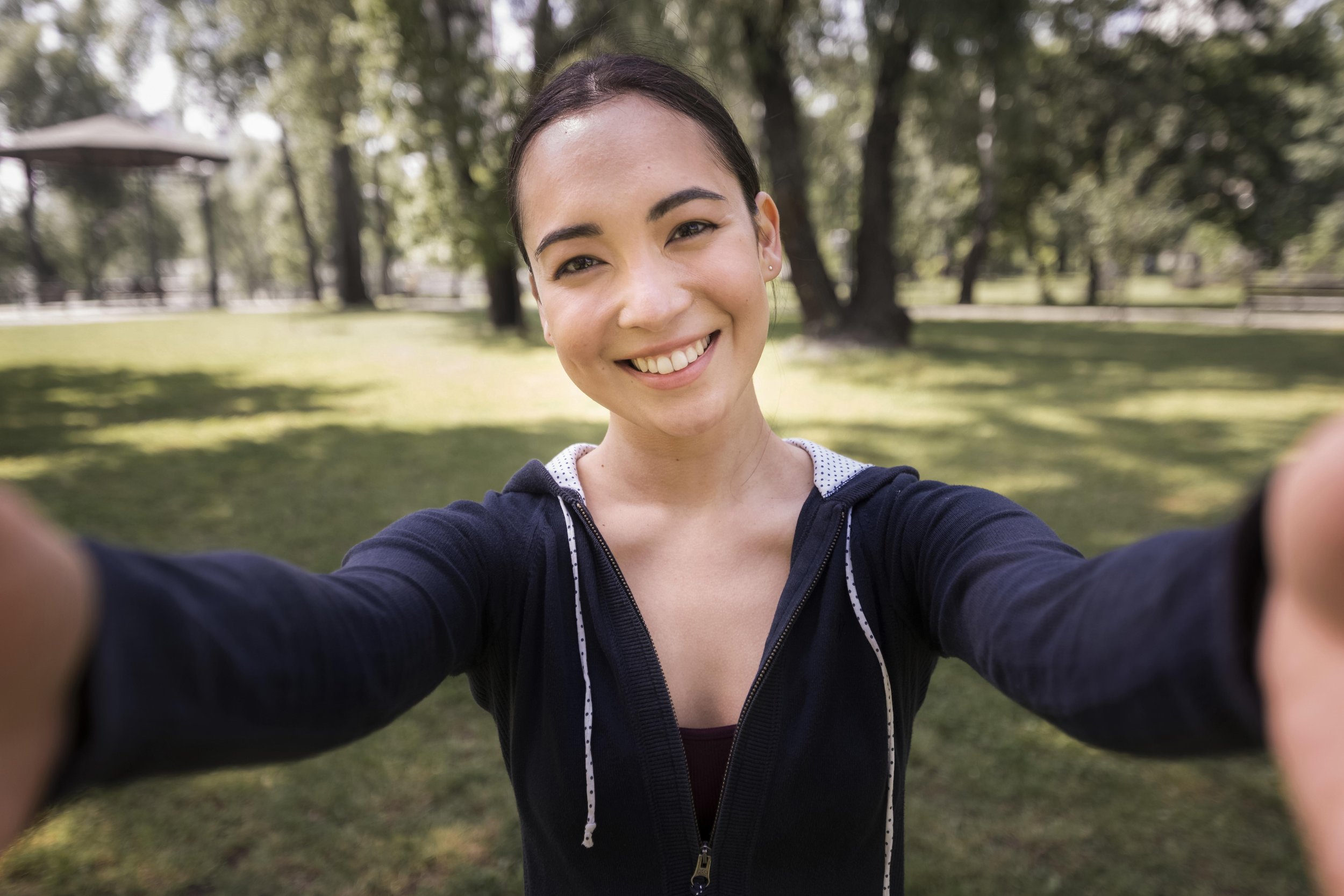 A woman taking a selfie outdoors in a park with trees and grass, smiling at the camera.