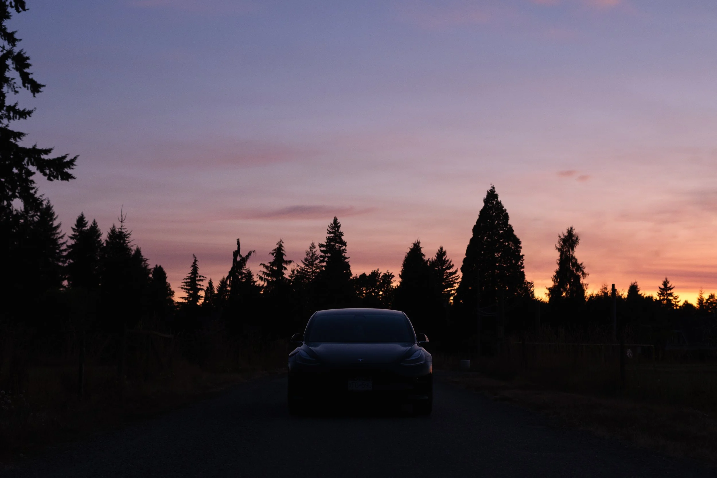 A car parked on a dark road at dusk with a colorful sunset sky and silhouetted trees in the background.