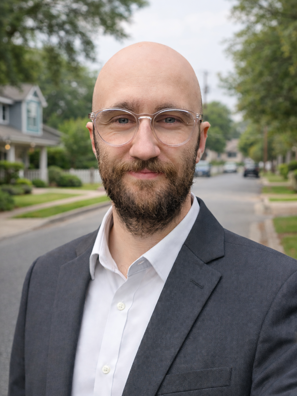 A man with a bald head, beard, and glasses smiling outdoors on a suburban street, wearing a dark suit and white shirt.
