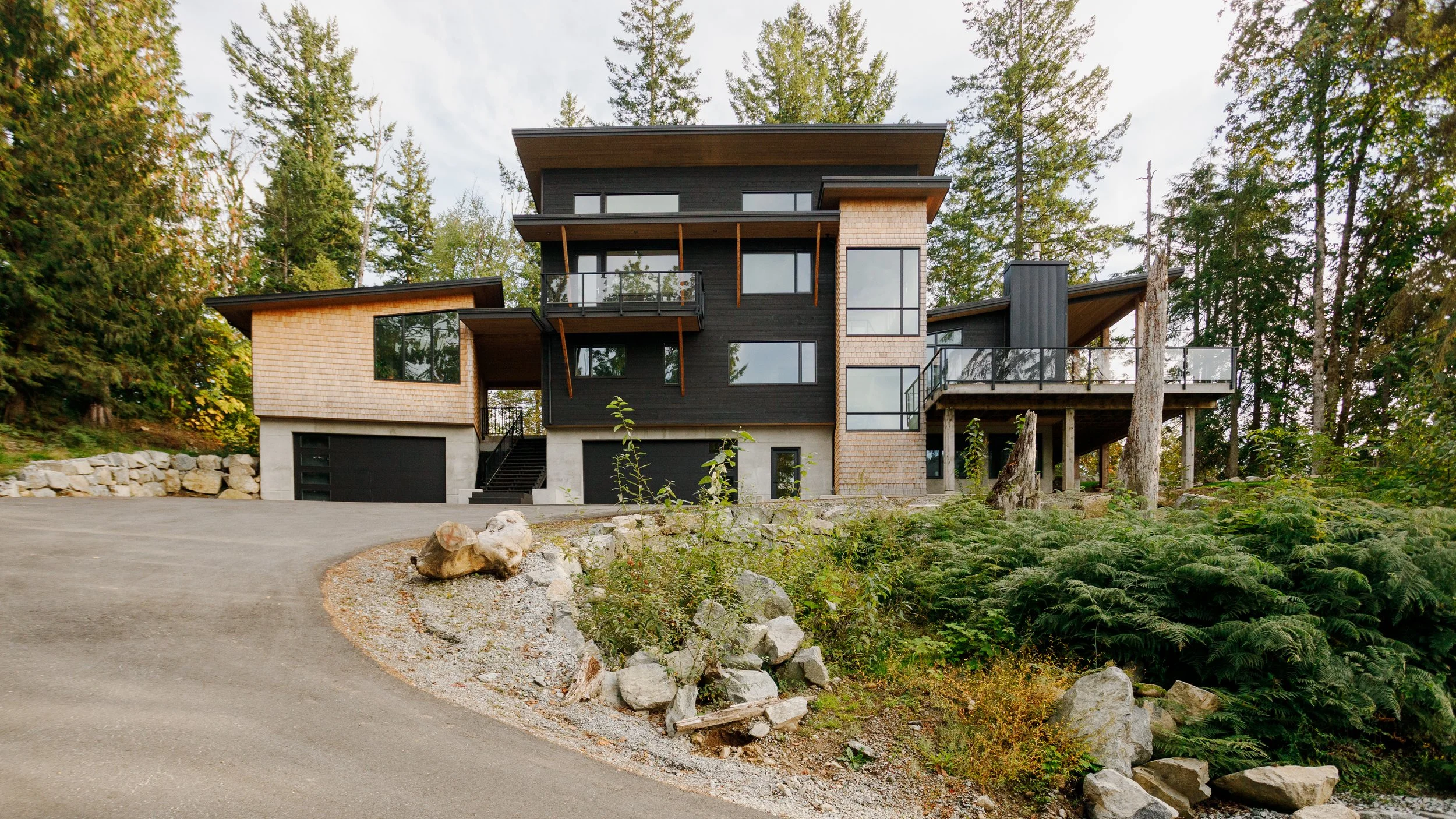Modern multi-story house with black and beige exterior, large windows, and multiple balconies surrounded by trees and greenery.