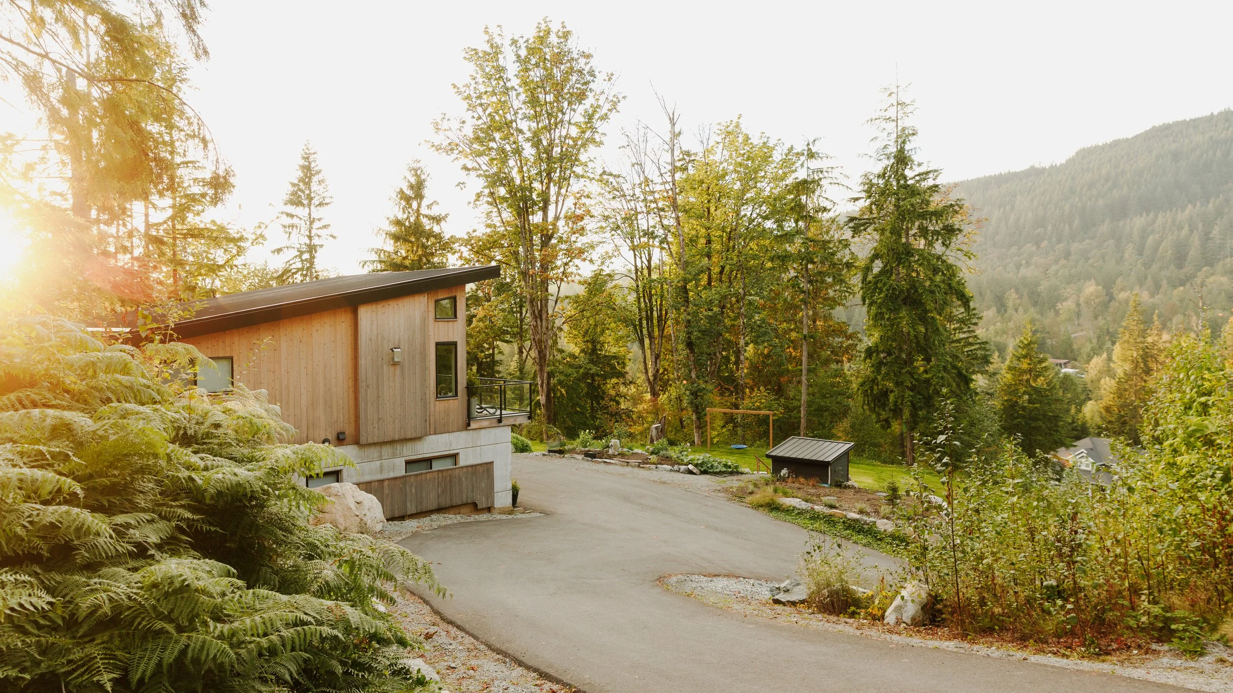 A modern house with wooden siding and a sloped roof, surrounded by trees and a mountainous landscape in the background, illuminated by golden sunlight.