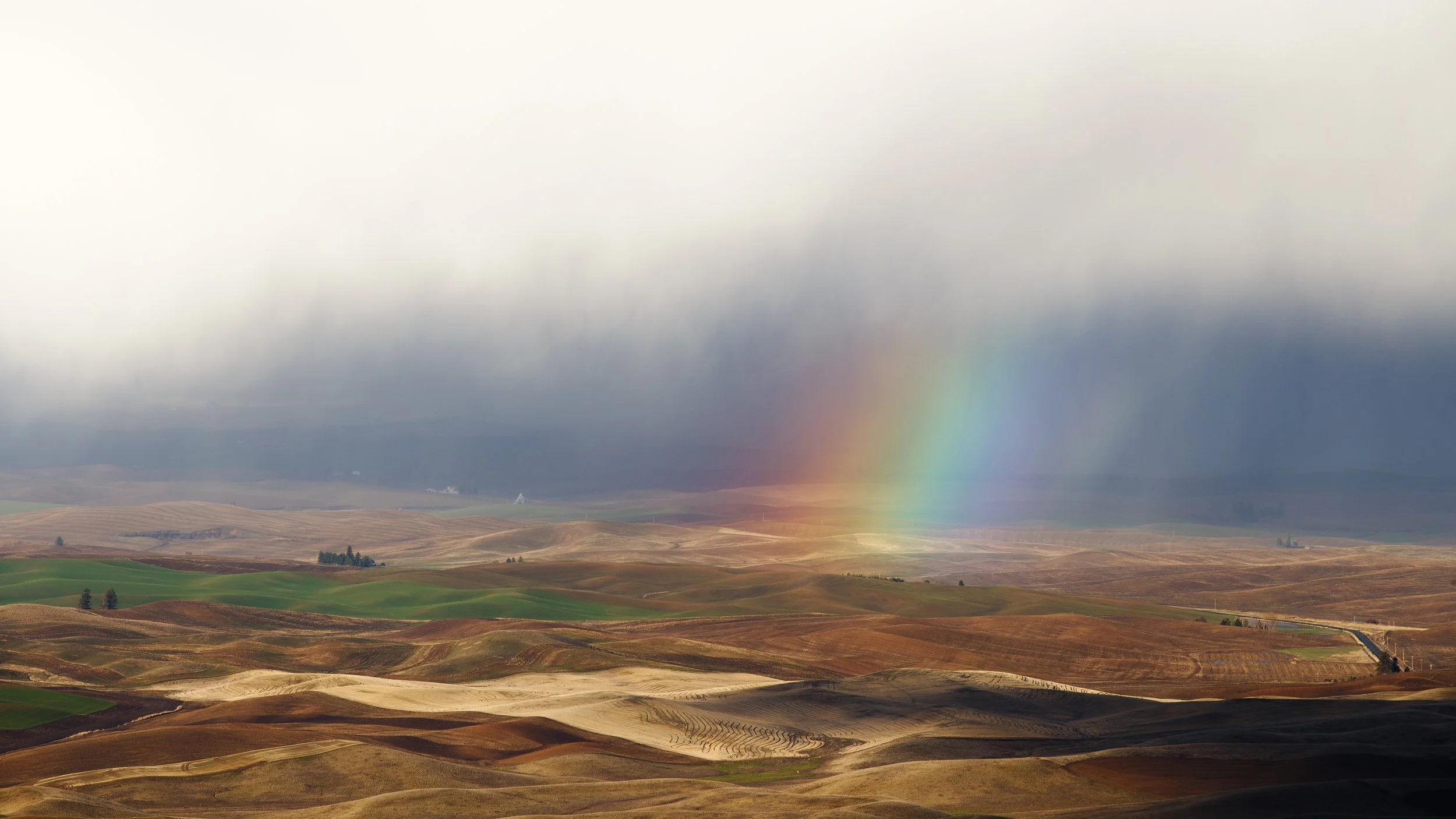 A vast landscape with rolling hills, partly covered with patches of green and brown fields, under a cloudy sky with a faint rainbow visible in the distance.