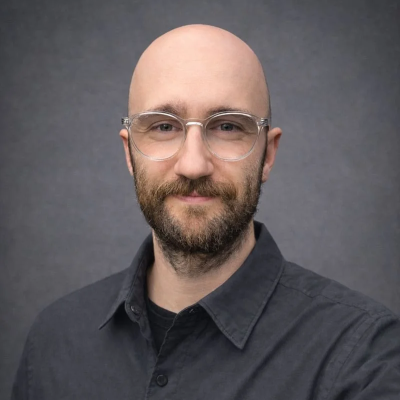 Portrait of a man with glasses and a beard, wearing a dark button-up shirt, against a gray background.