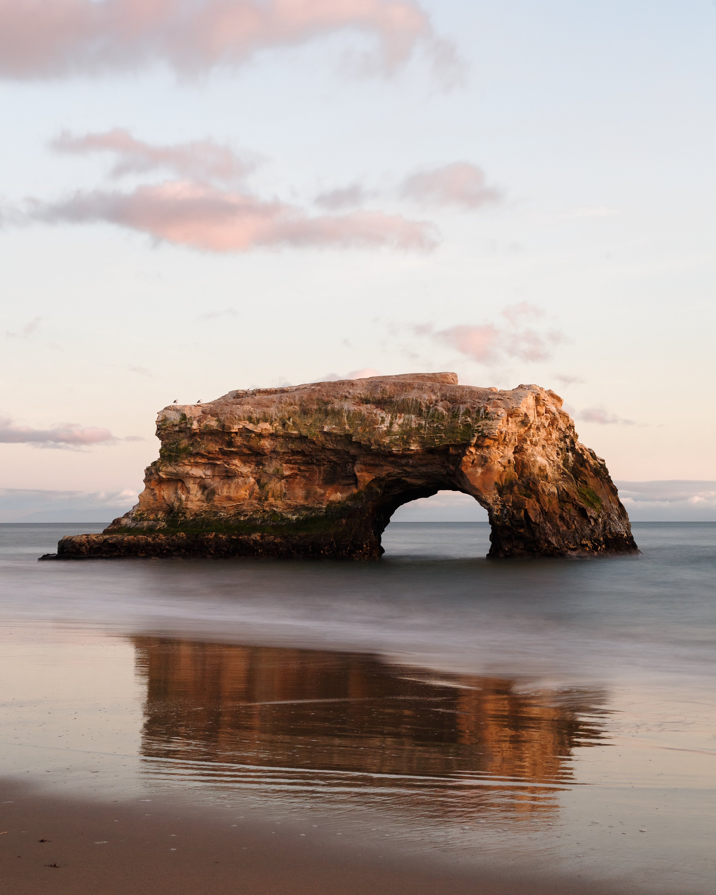 A coastal scene with a large rock formation featuring a natural arch, reflected in the water, under a sky with scattered clouds at sunset.