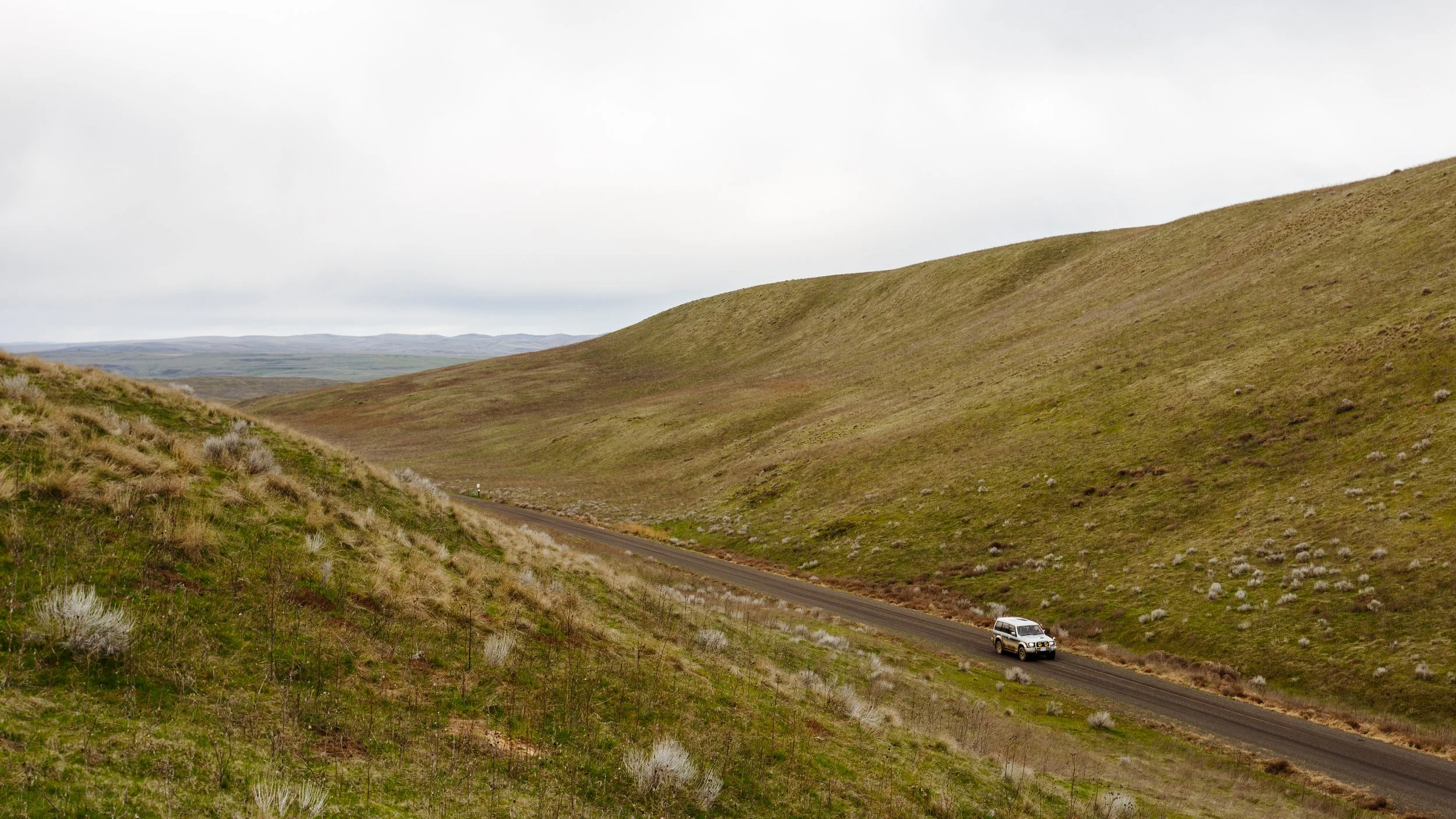 A car on a narrow highway winding through rolling grassy hills under a cloudy sky.