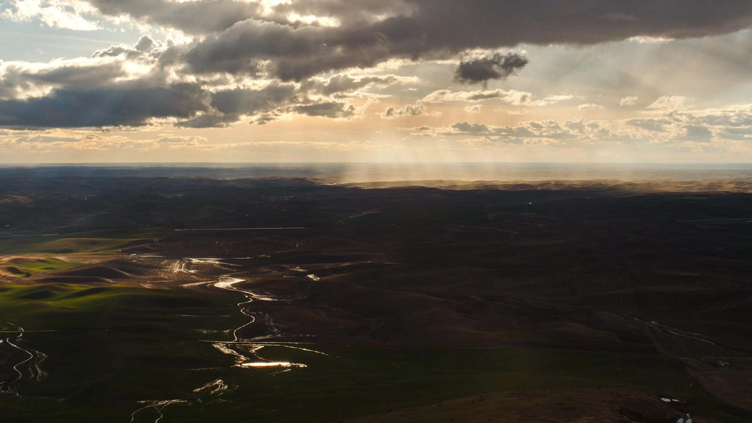 Landscape view of rolling hills and fields at sunset, with clouds overhead and sunlight breaking through, casting reflections on wet ground and water channels.