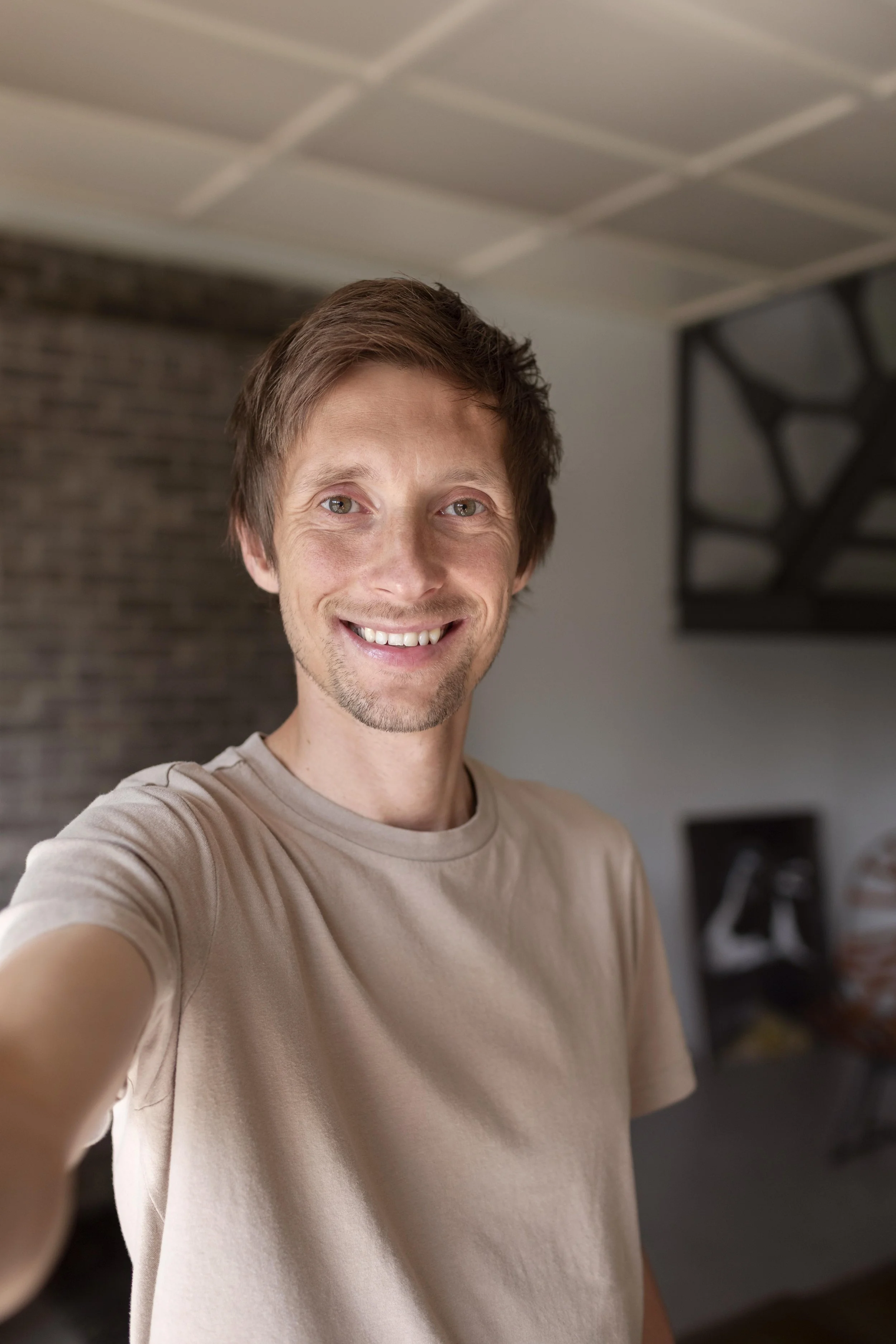 A smiling man taking a selfie indoors, with a modern interior design featuring brick wall and artistic wall decor.