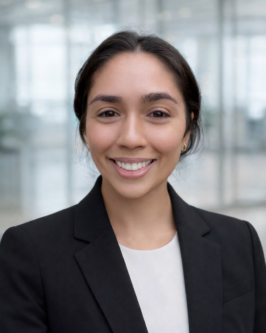 A young woman with dark hair, smiling, wearing a black blazer and a white top, in an office setting with glass windows.