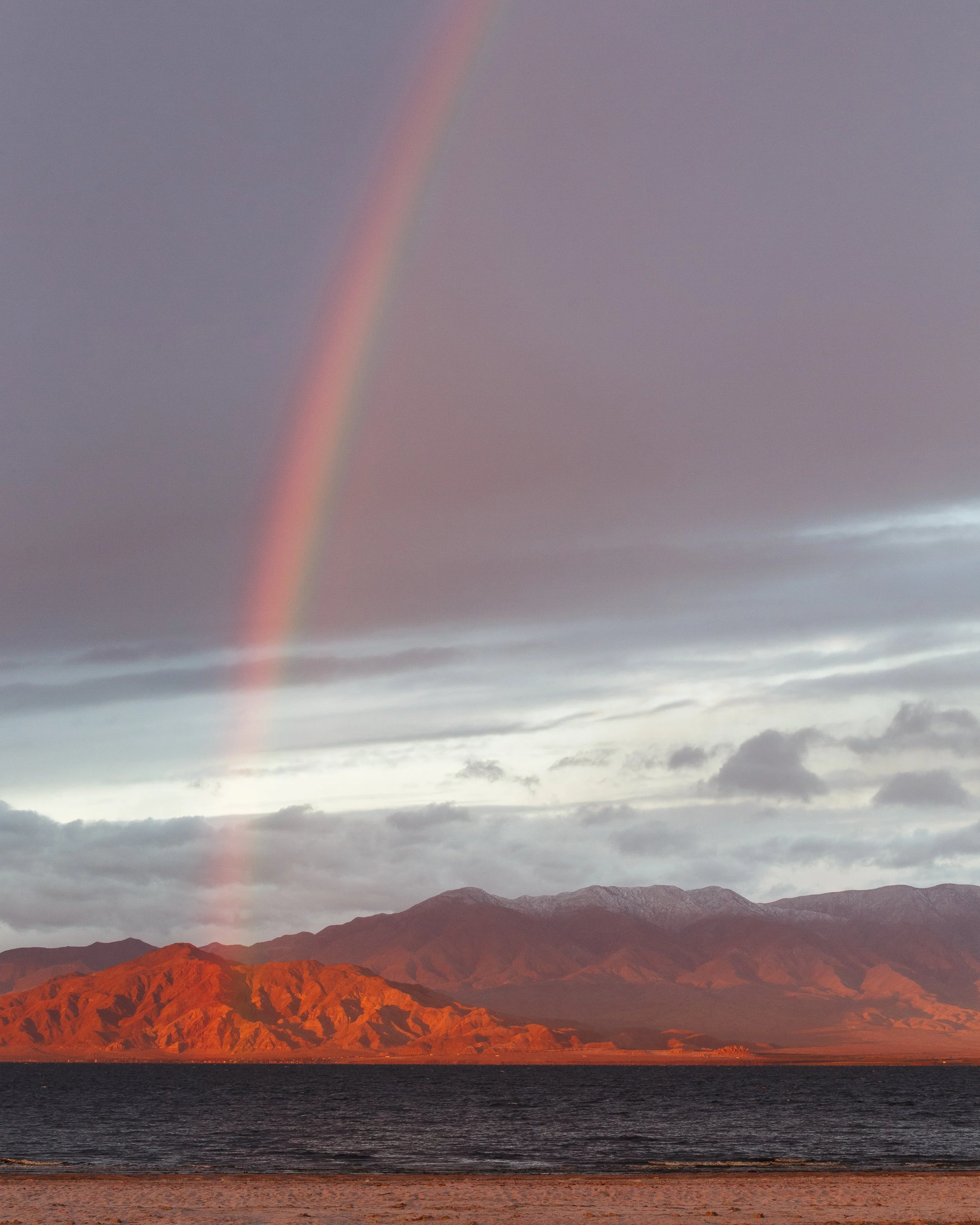 Rainbow arcing over a mountain range with a dark cloudy sky above and a body of water and sandy beach in the foreground.