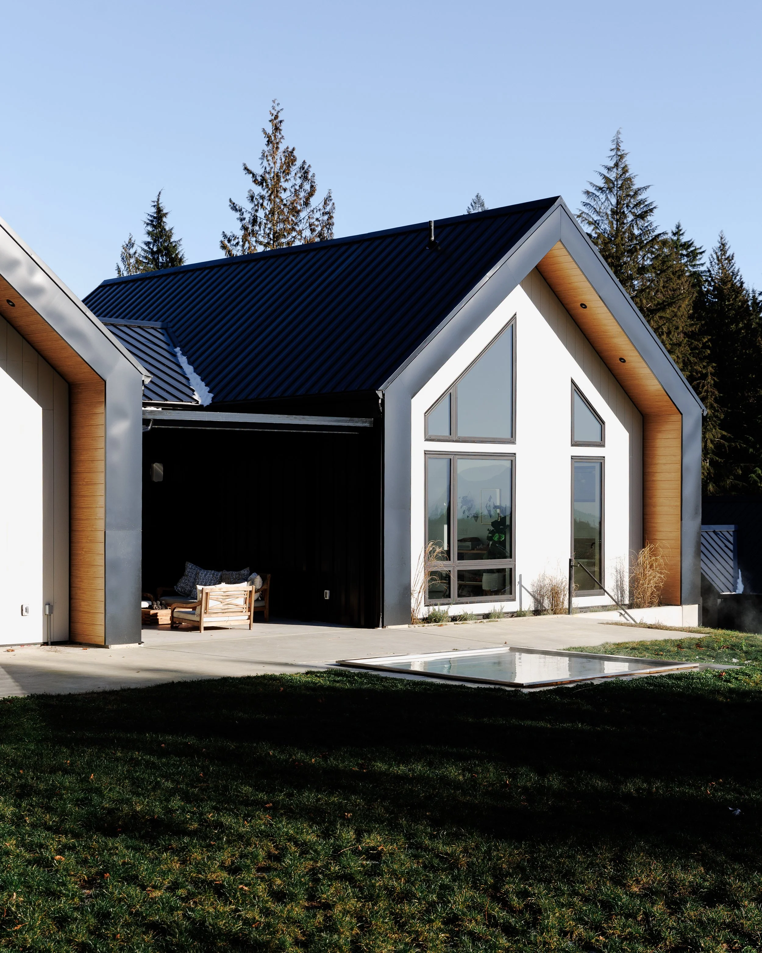 Modern house with a steep black metal roof, large triangular windows, and wooden accents, set against a background of tall trees and a clear blue sky.