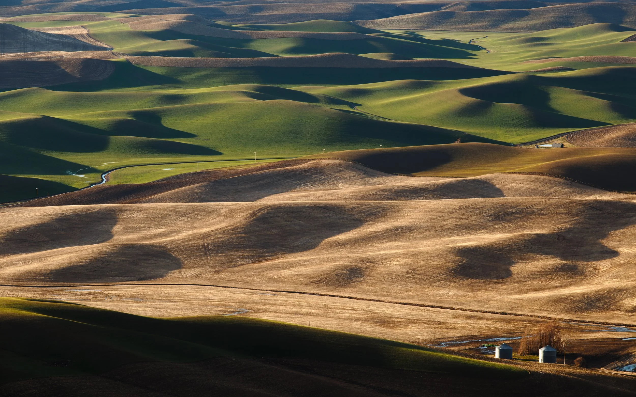 Rolling hills with patches of green and brown fields, some with shadows, and two silos at the bottom right.