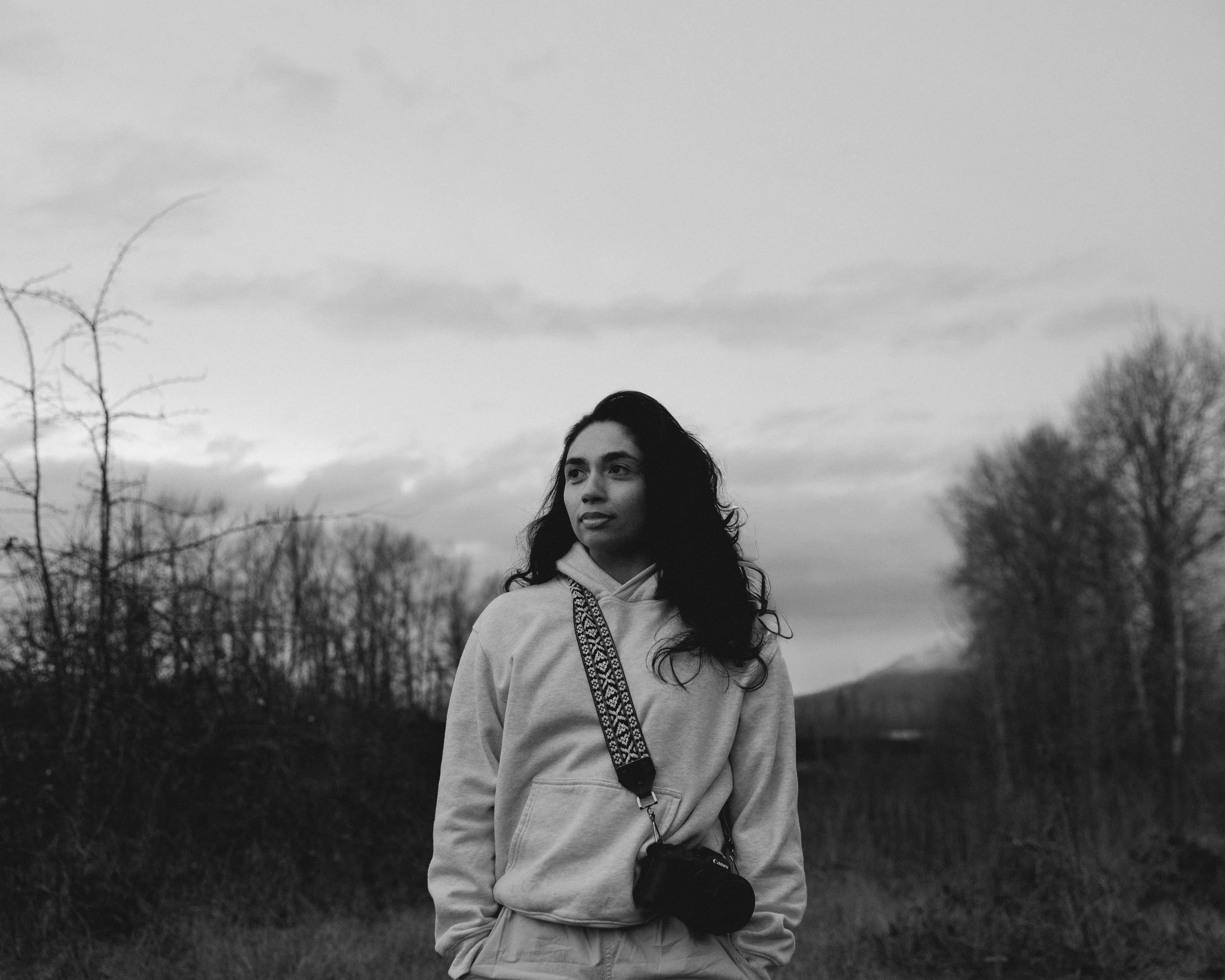 A woman with dark, wavy hair wearing a light-colored hoodie and carrying a camera with a patterned strap standing outdoors in a natural setting with trees and cloudy sky in the background