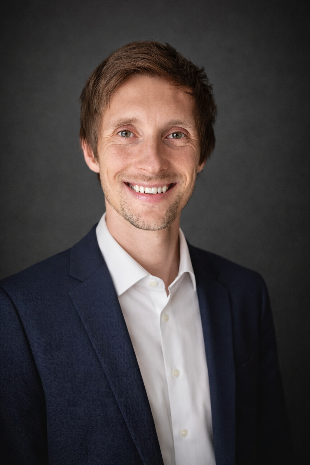 Headshot of a young man with brown hair, green eyes, smiling, wearing a white shirt and navy blazer, against a dark gray background.