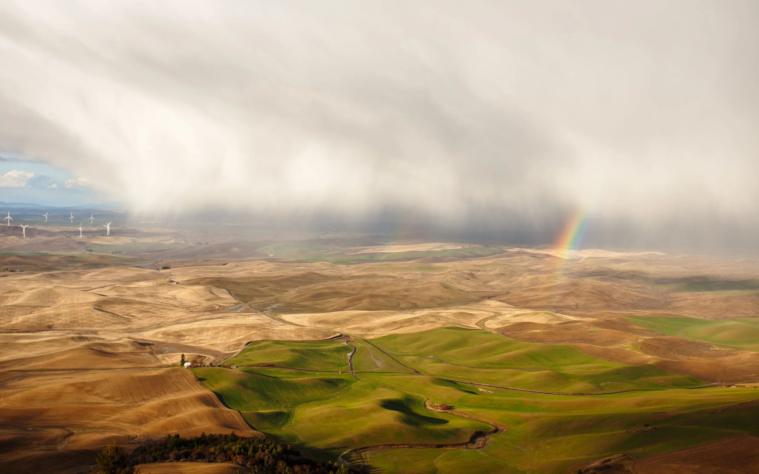 Scenic view of rolling hills with brown and green fields, a rainbow in the cloudy sky, and wind turbines in the distance.
