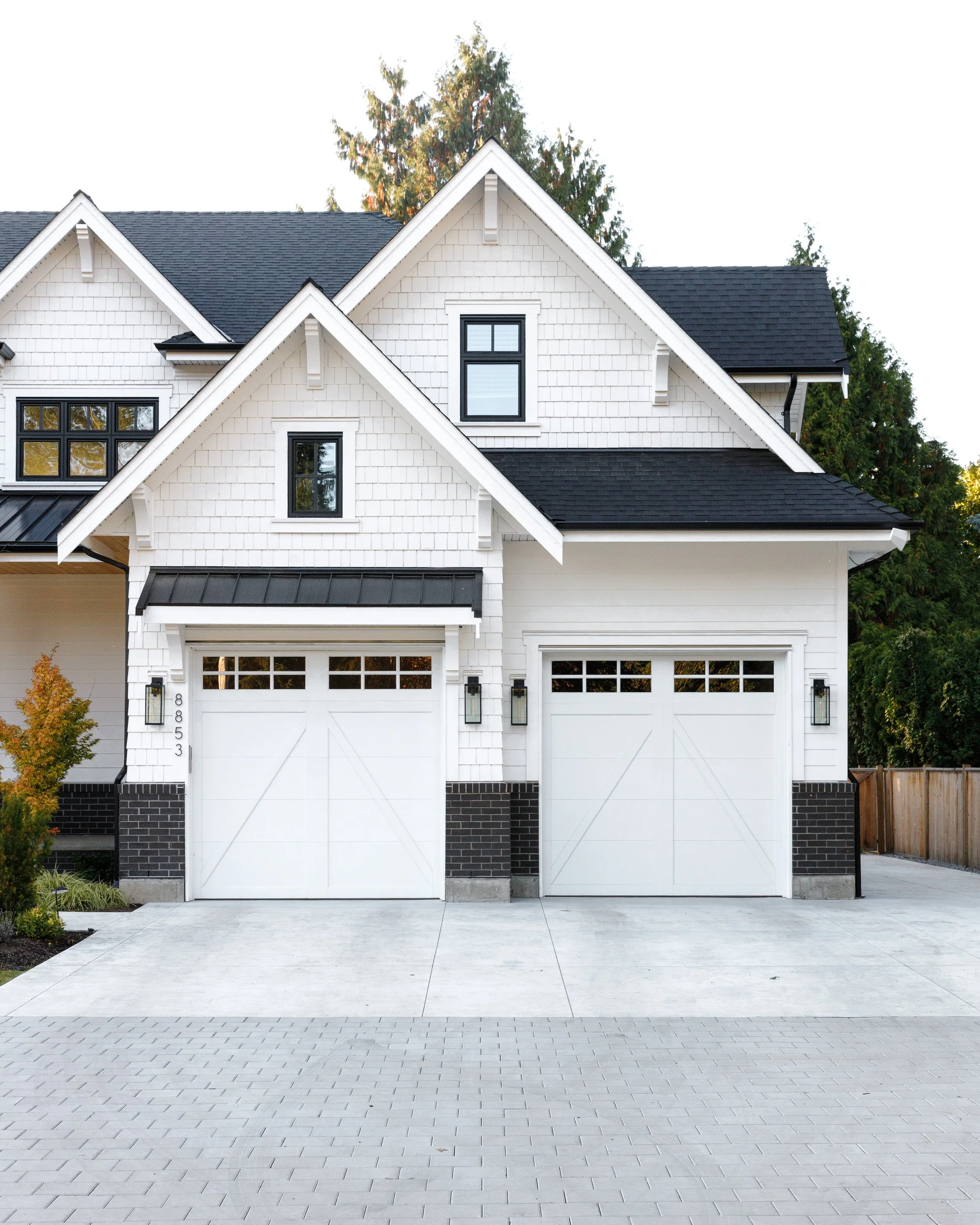 Modern white house with attached two-car garage and black roof, set in a clean paved driveway with some greenery and trees in the background.