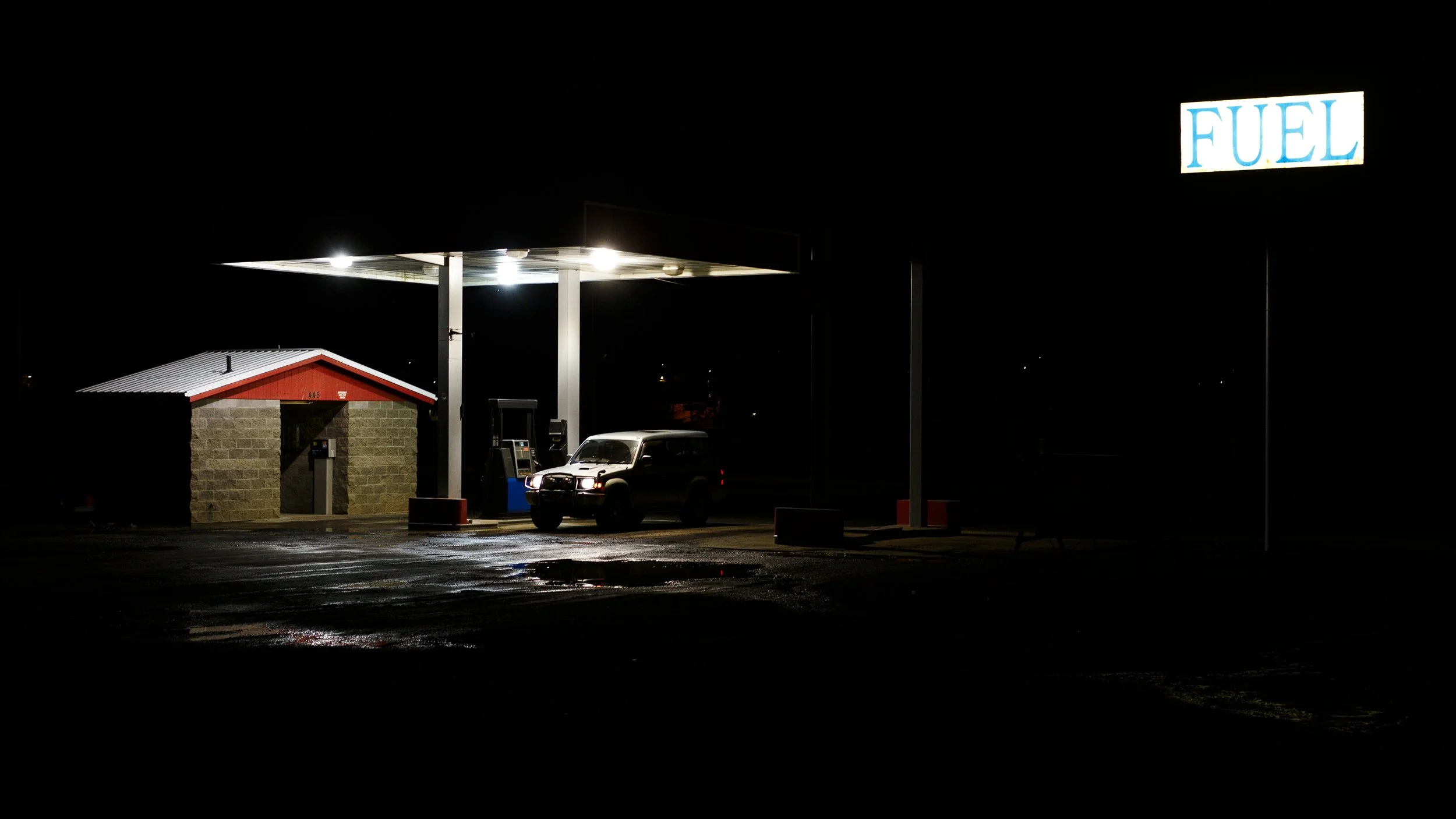 Empty gas station at night with a single car parked under the bright canopy and a illuminated 'FUEL' sign.