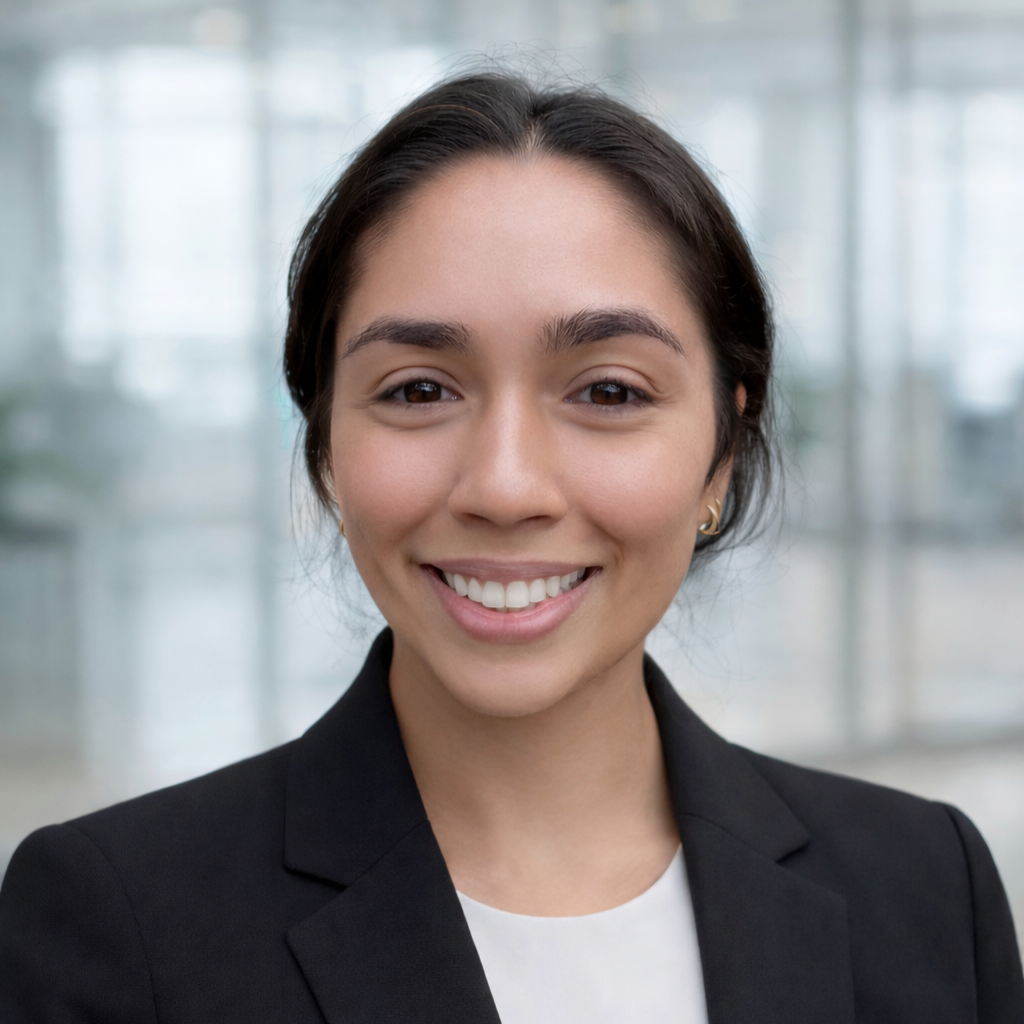 A young woman with dark hair, wearing a black blazer and a white shirt, smiling indoors with a blurred background of glass windows and natural light.