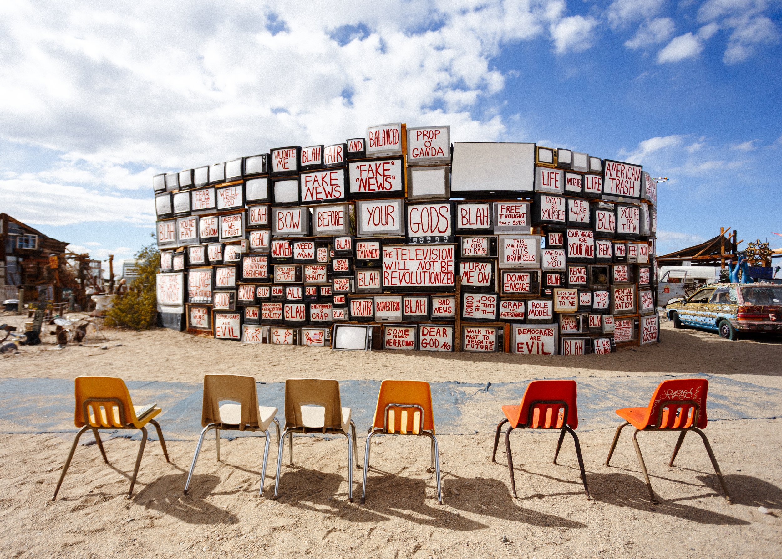 An outdoor art installation featuring stacked vintage TVs with red and white signs displaying messages and slogans, with five colorful chairs in the foreground and a partly cloudy sky overhead.