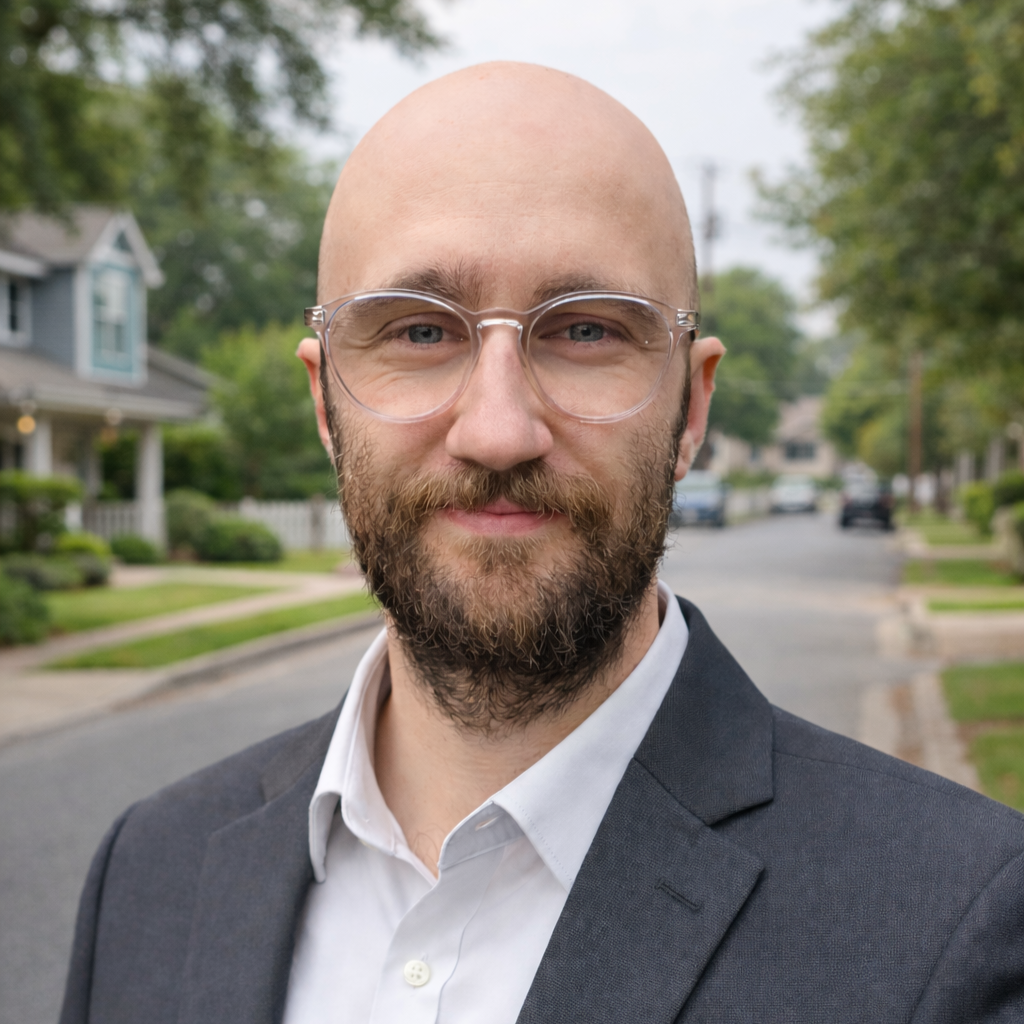 A man with glasses and a beard wearing a suit jacket over a white shirt, standing outdoors on a residential street lined with houses and trees.