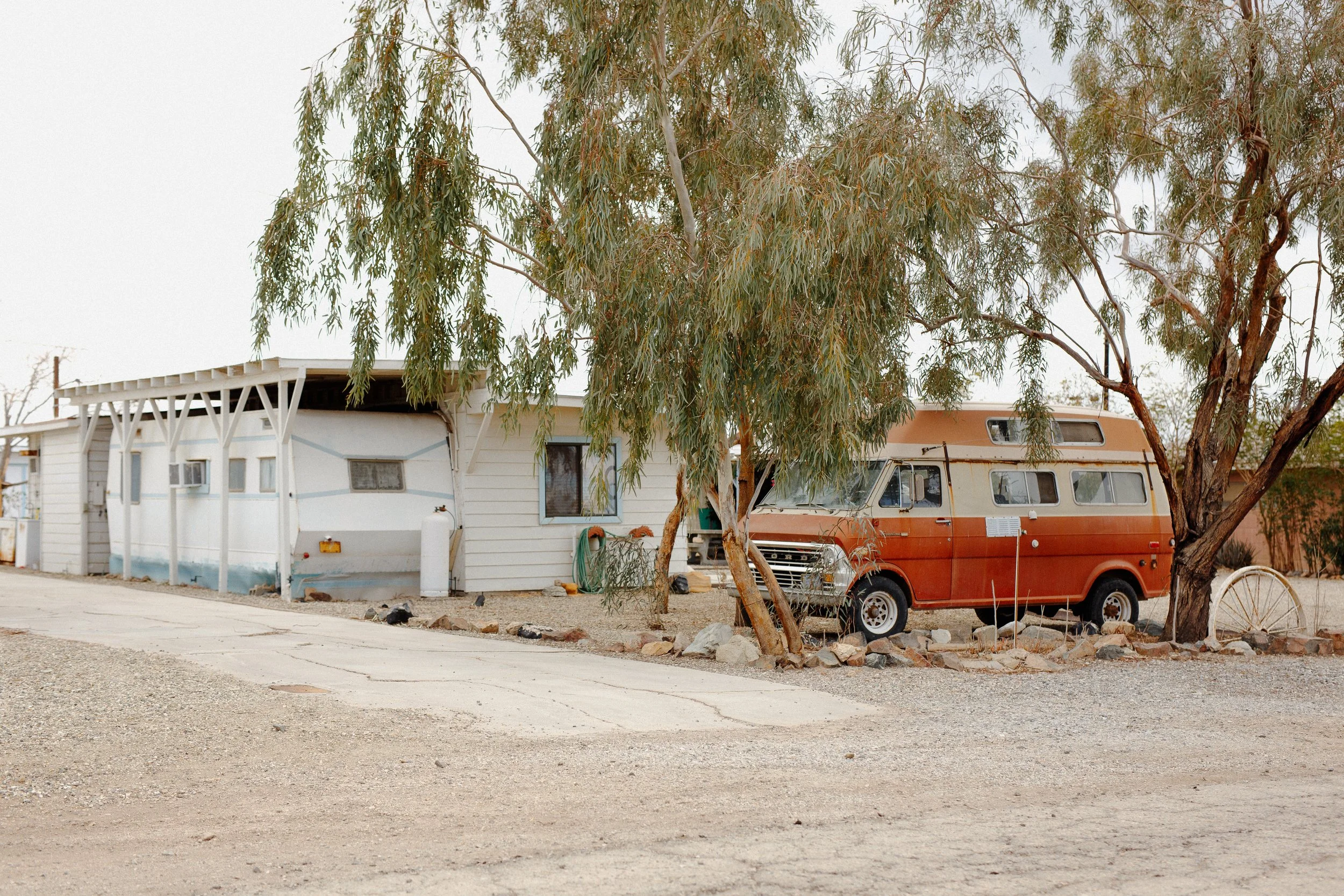 A mobile home with a white exterior, a covered porch with white supports, and a propane tank in front. An orange and white vintage camper van parked beside a tree with sparse green leaves. Gravel ground and a distant wooden fence in a desert landscap