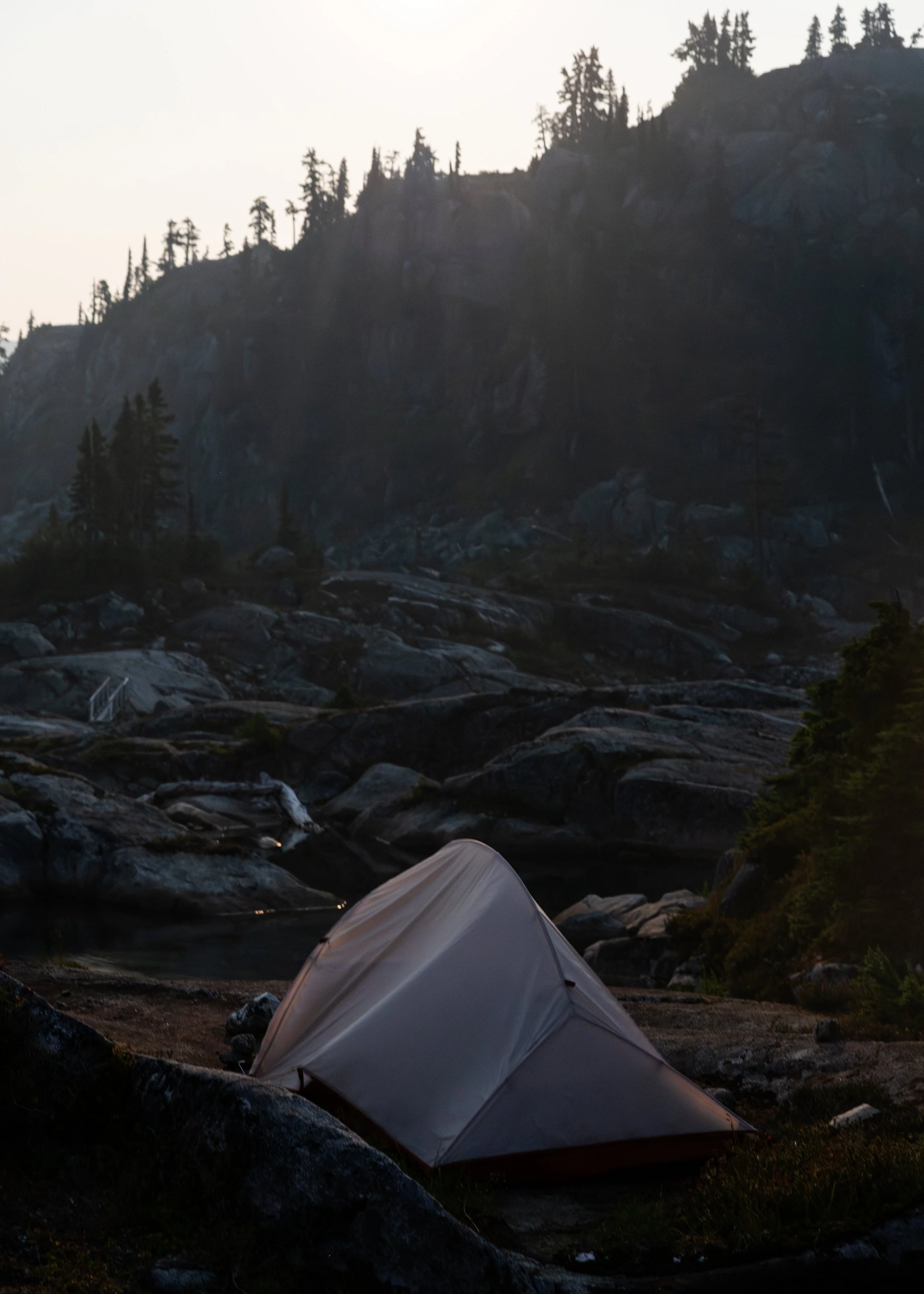 A white tent set up beside a rocky river with a mountain and trees in the background, likely during dusk or dawn.