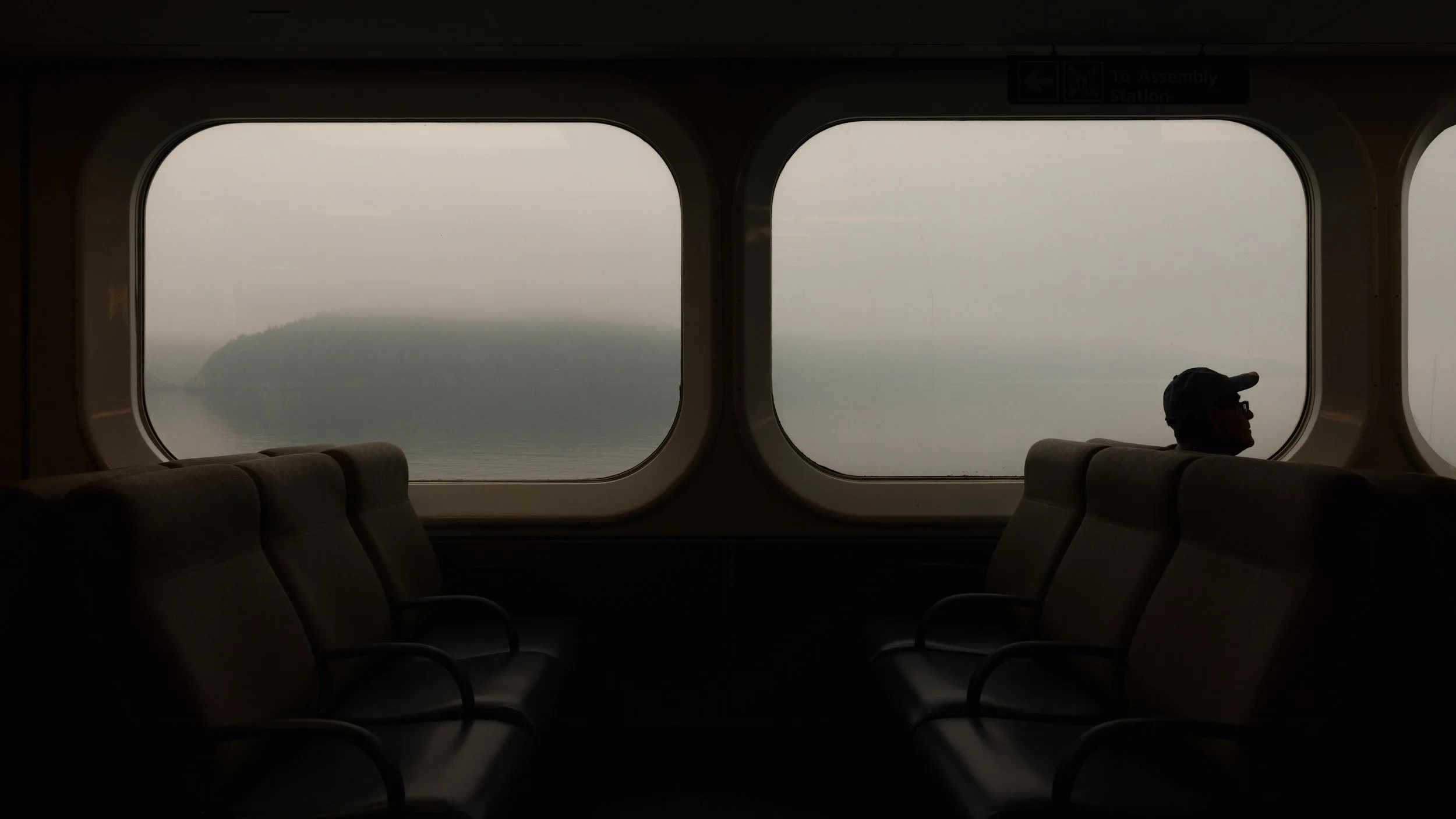 Interior of a ferry with empty seats and a silhouette of a person with a cap looking out the window at foggy water and a distant landmass.