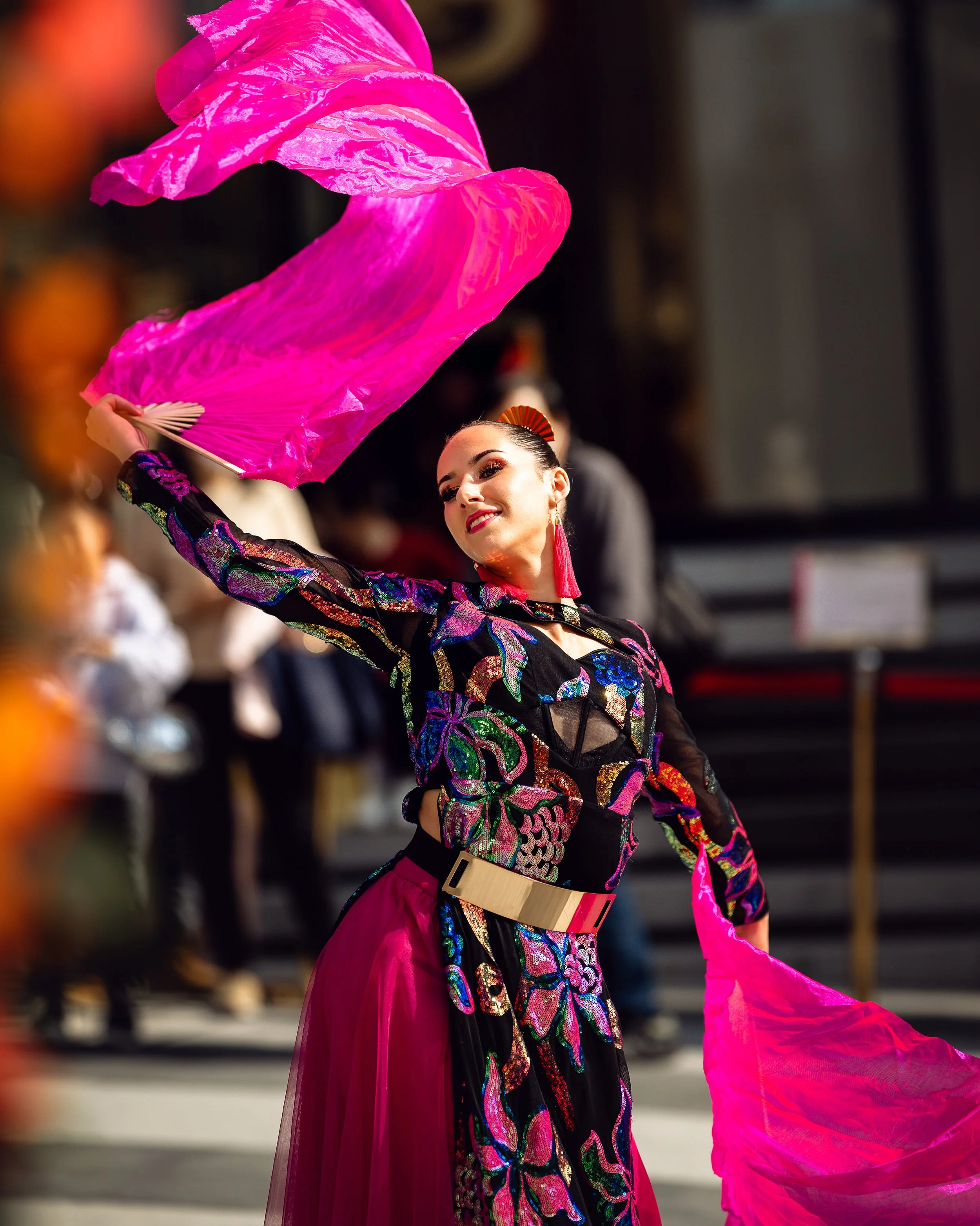 A close-up of a BPM Productions Hong Kong performer in a colorful, sequined outfit gracefully handling a vibrant pink silk fan during an outdoor show in Macau.
