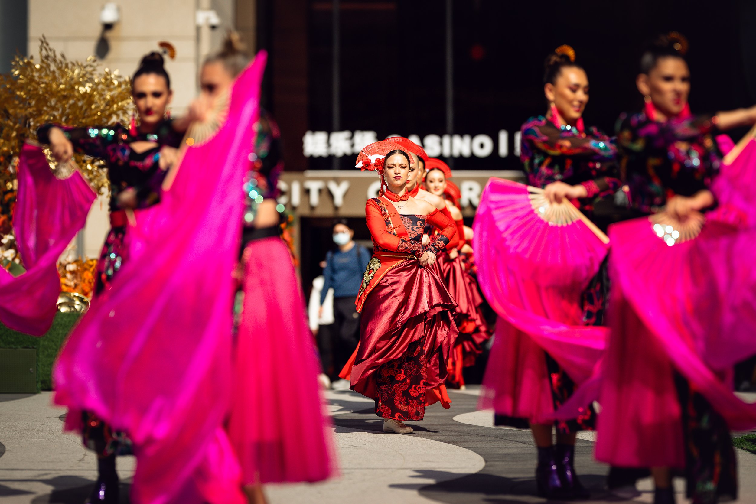 A synchronized group dance routine by BPM Productions Hong Kong performers with large pink fans in front of the City of Dreams casino entrance.