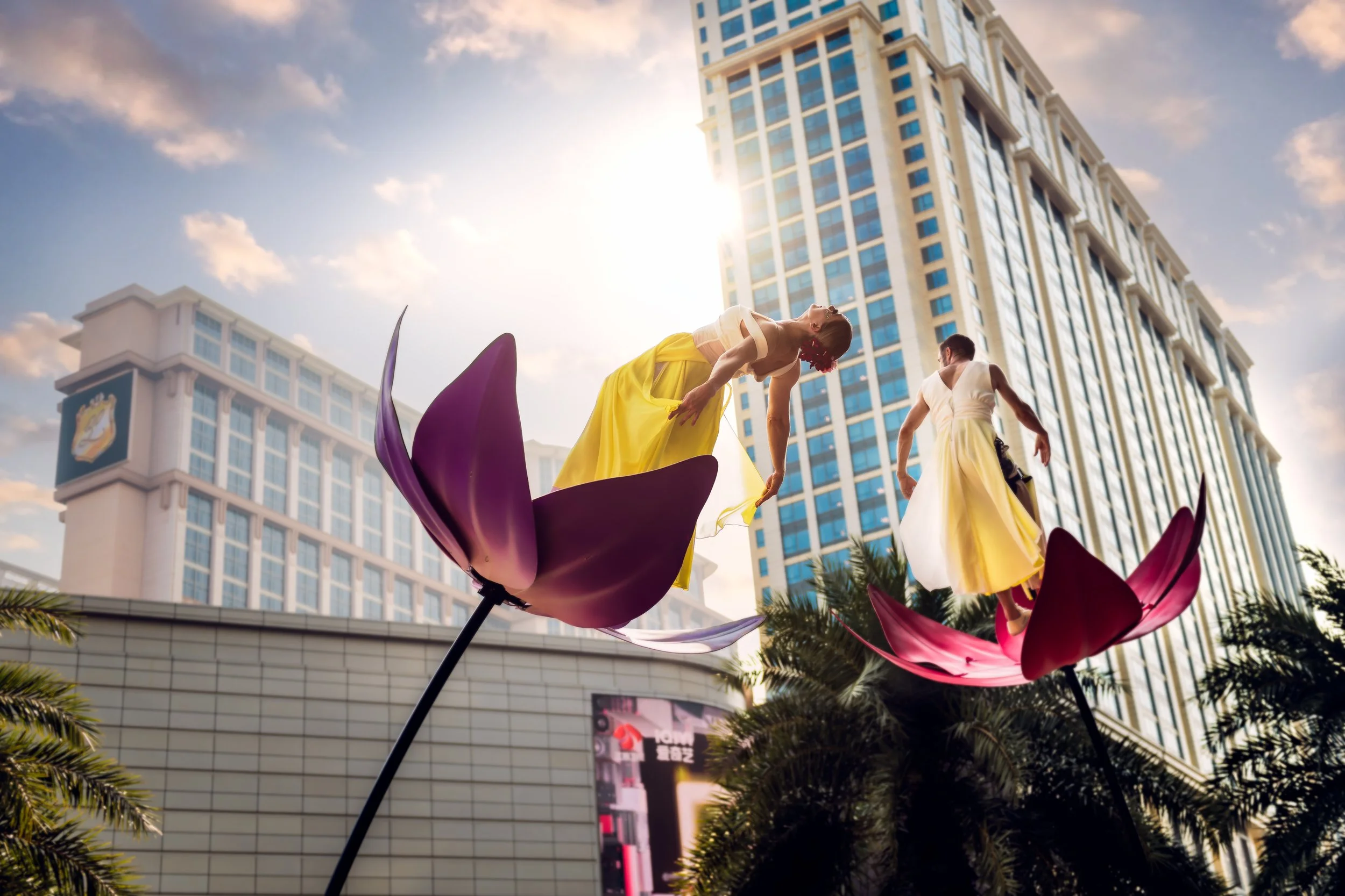 Two acrobatic dancers from BPM Productions Hong Kong performing on sway poles above large pink and yellow flower props during a sunset outdoor activation.