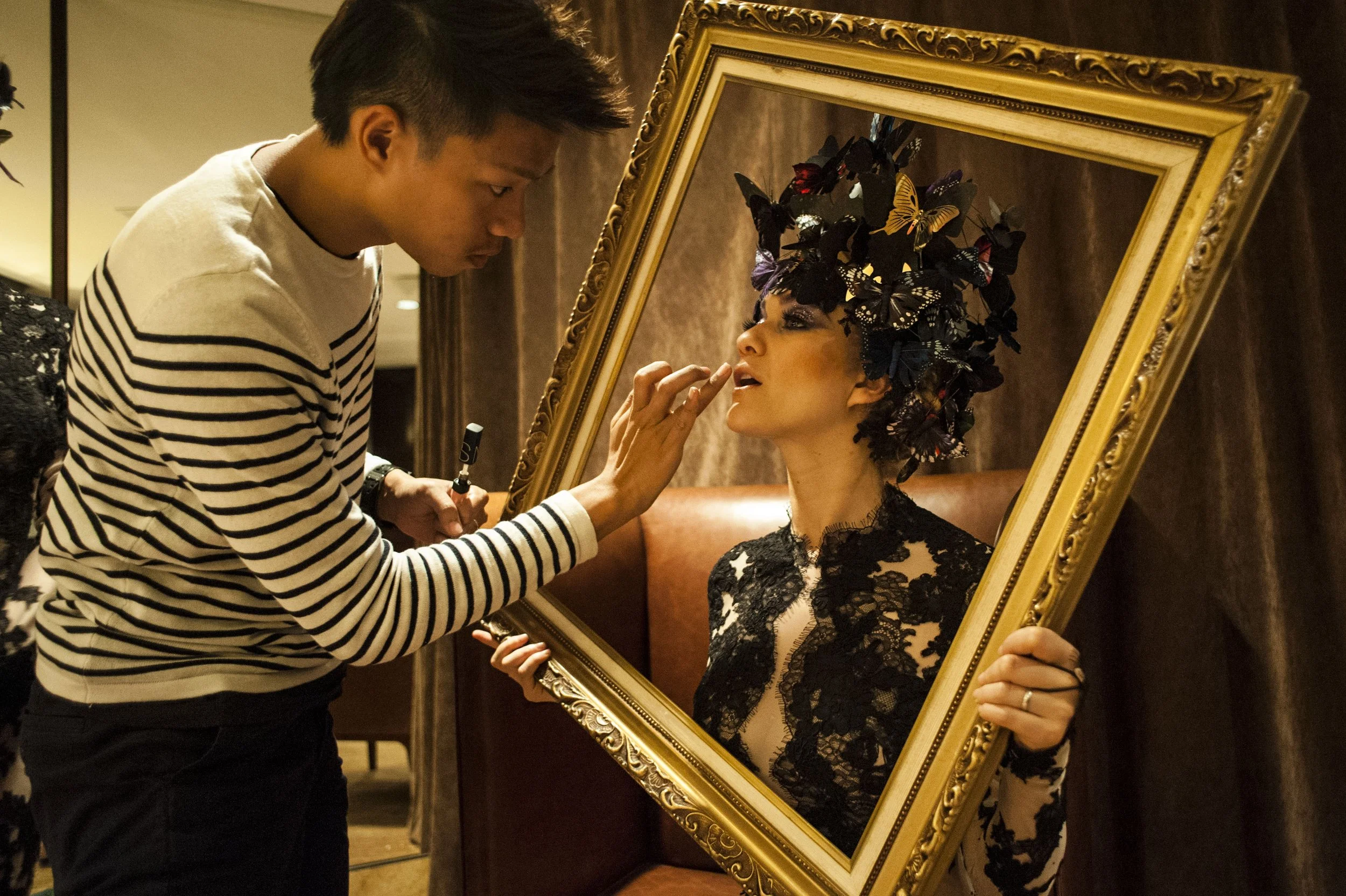Behind-the-scenes shot of a makeup artist refining a performer's look for a Clé de Peau Beauté event, featuring a golden frame and intricate floral headpiece.