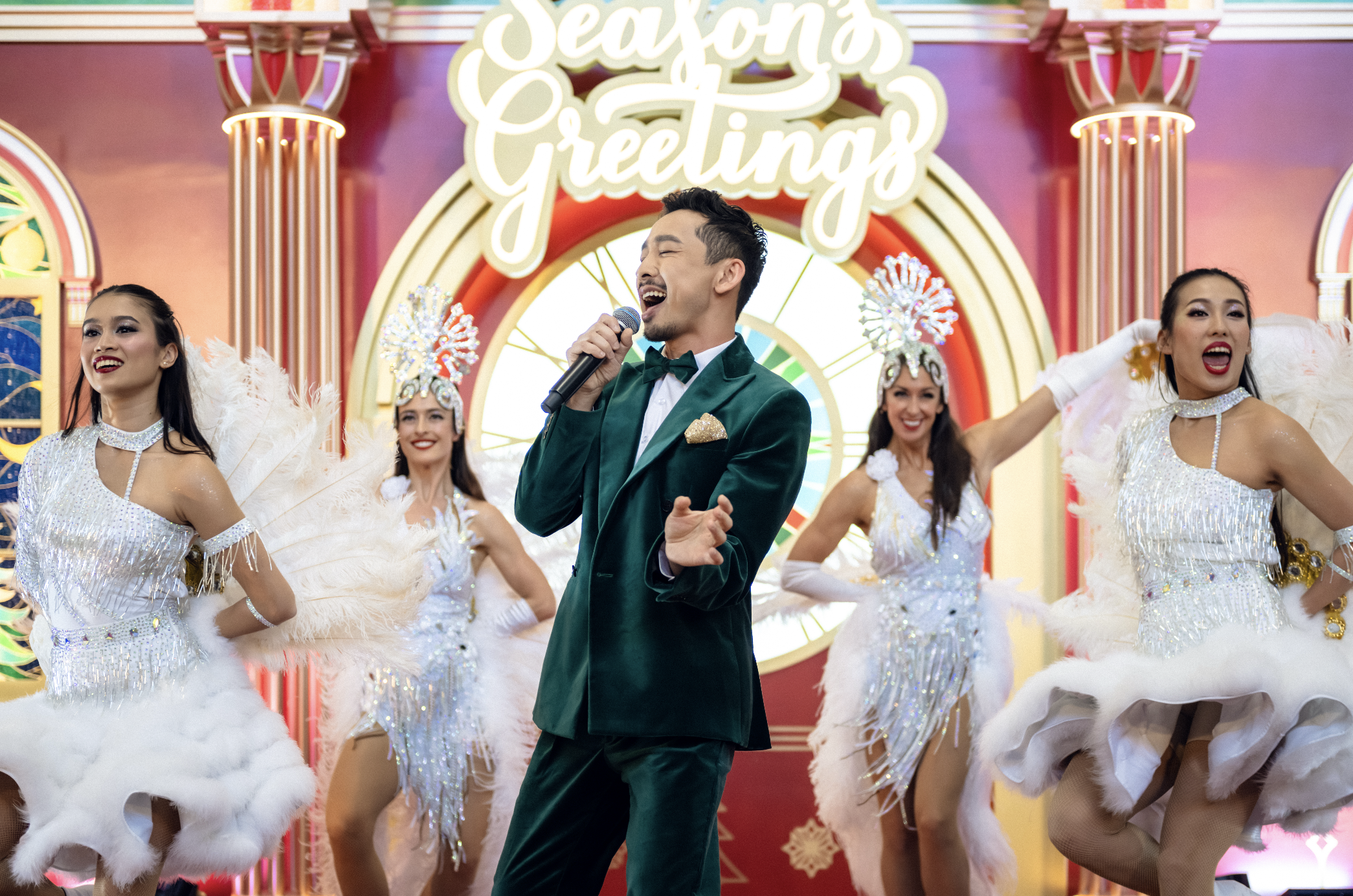 A male lead singer and troupe of dancers from BPM Productions Hong Kong perform a Broadway-style Christmas show in front of a "Season's Greetings" backdrop at Hong Kong International Airport Terminal 1.