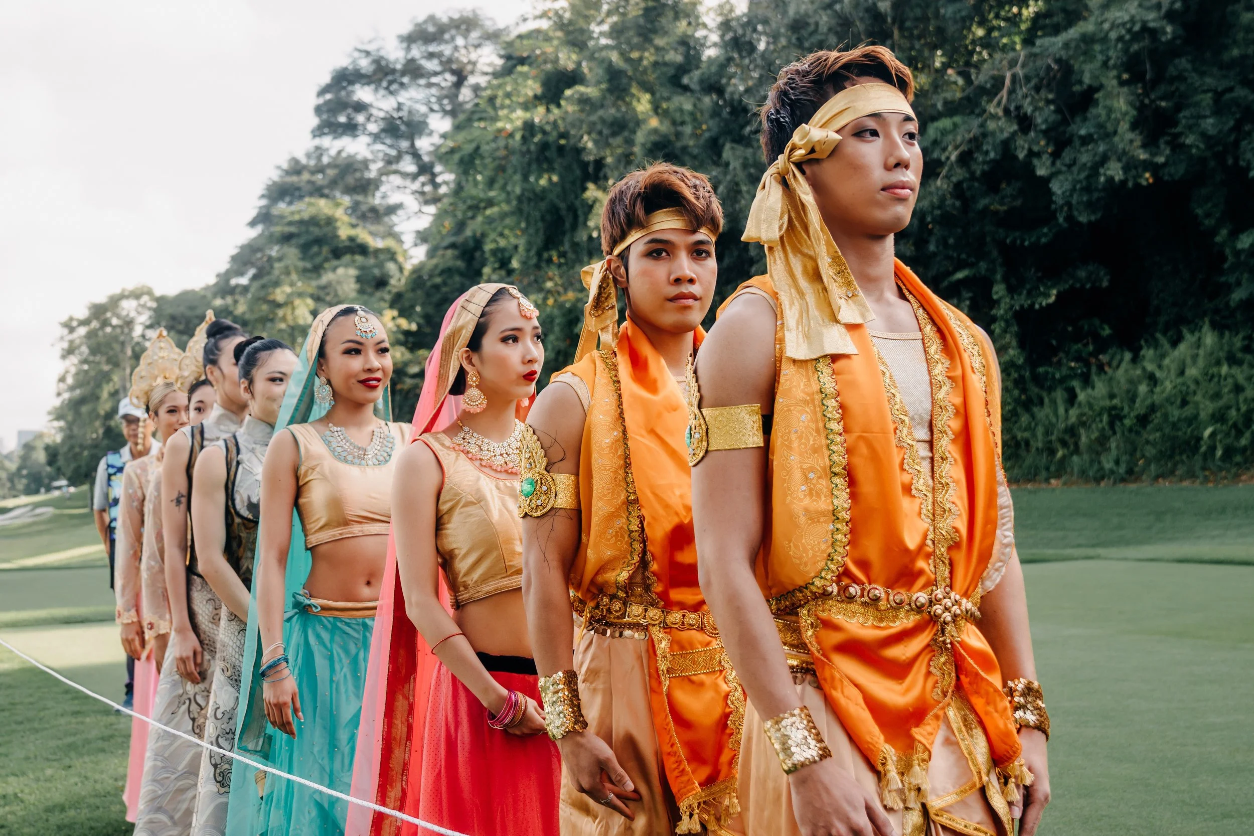 Performers dressed in ornate, colorful traditional attire, including gold headbands and silk fabrics, stand in a line during the LIV Golf opening show in Singapore.