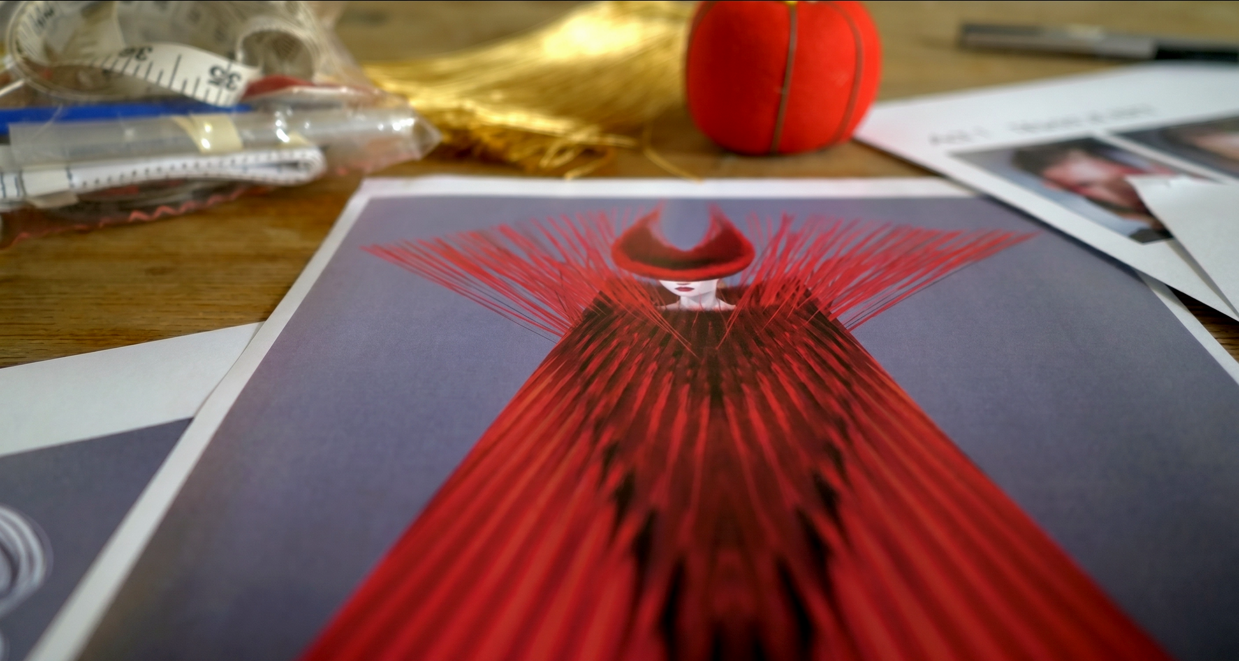 A printed fashion illustration of a woman in a red dress with exaggerated red lines, with a red hat covering her eyes, on a desk among papers, a red round object, a plastic bag with measuring tape, and a pen.