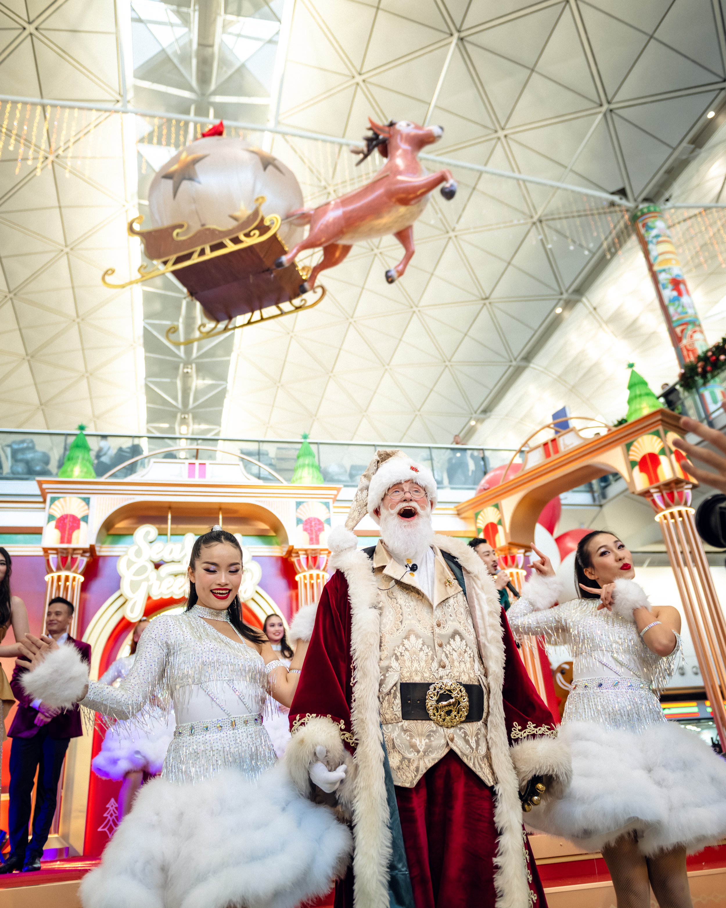 Santa Claus and festive dancers from BPM Productions Hong Kong perform in front of a "Season's Greetings" stage with a flying reindeer display overhead at Hong Kong International Airport.