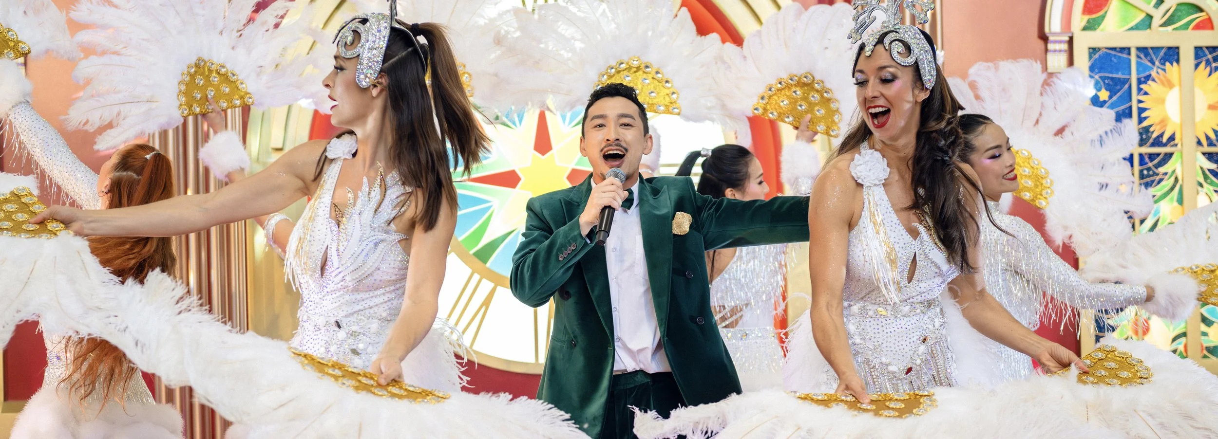 A male singer in a green velvet blazer performs alongside dancers in white feathered costumes for a Christmas musical show at Hong Kong International Airport.