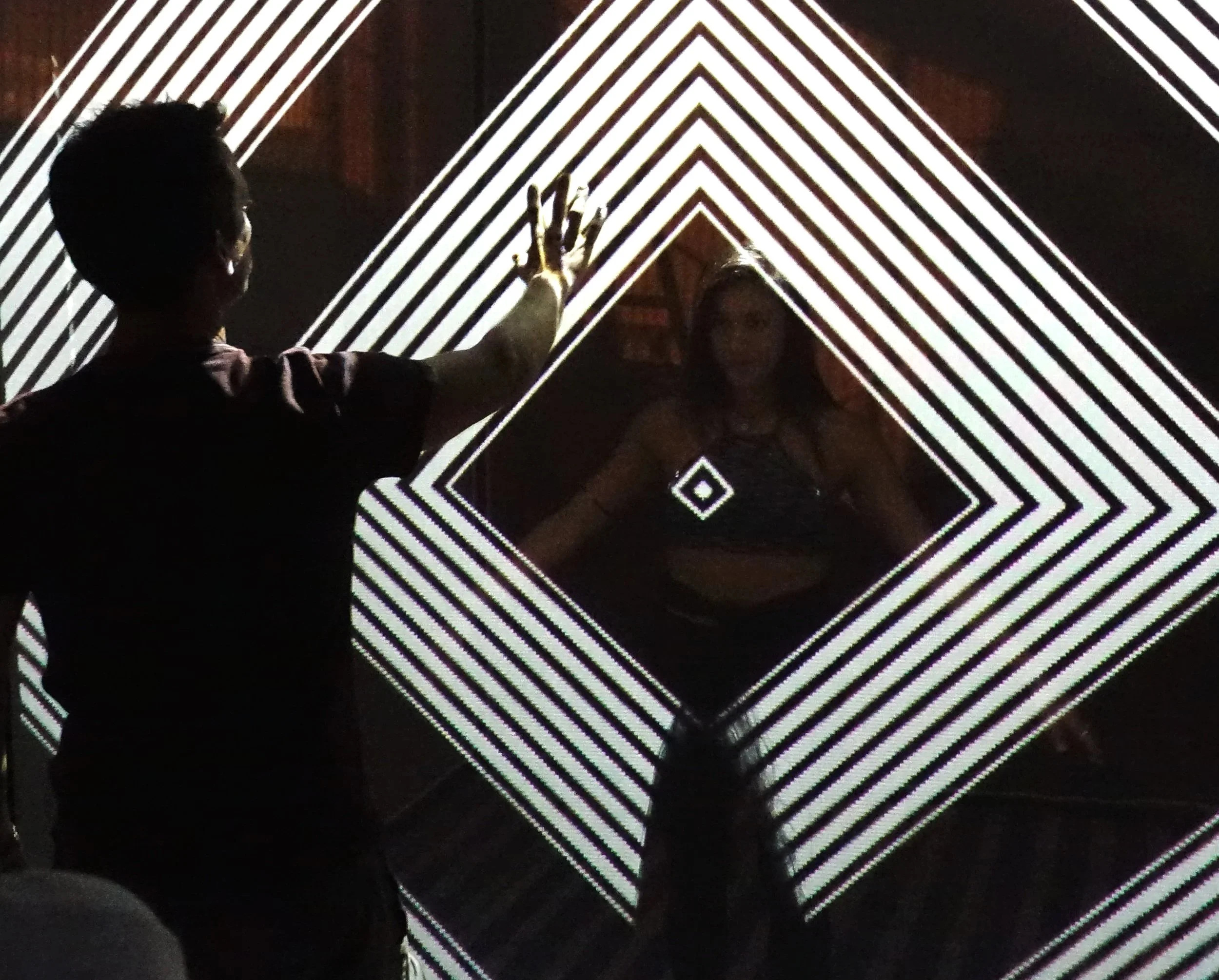 A person with short hair and dark skin reaching out with their right hand to touch a digital display with black and white geometric patterns and neon green accents on a dark background.